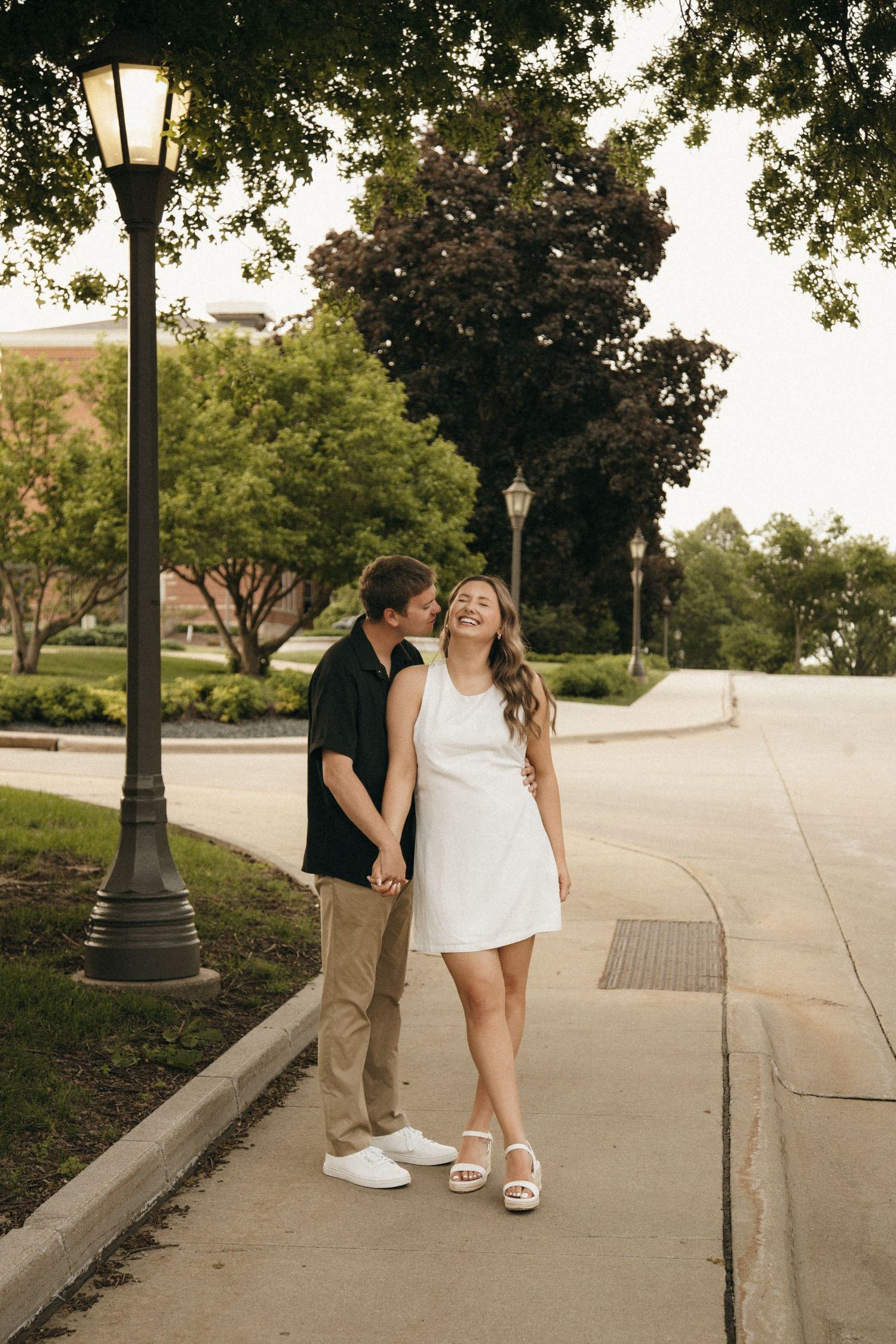 Happy couple walking hand-in-hand on a tree-lined path, sharing a tender kiss near a vintage street lamp at dusk.