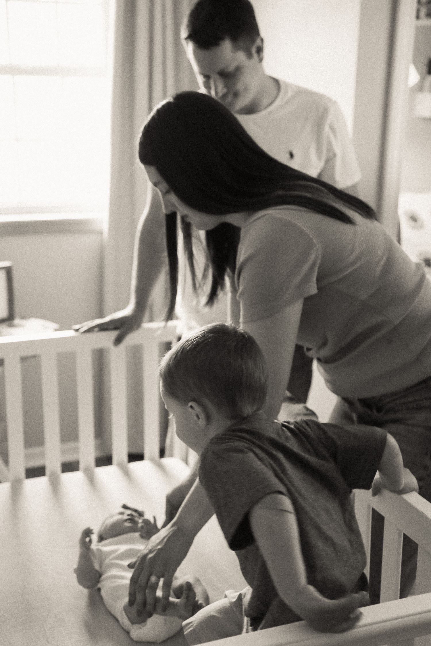 Parents lean over a white crib watching their baby in a naturally lit nursery.