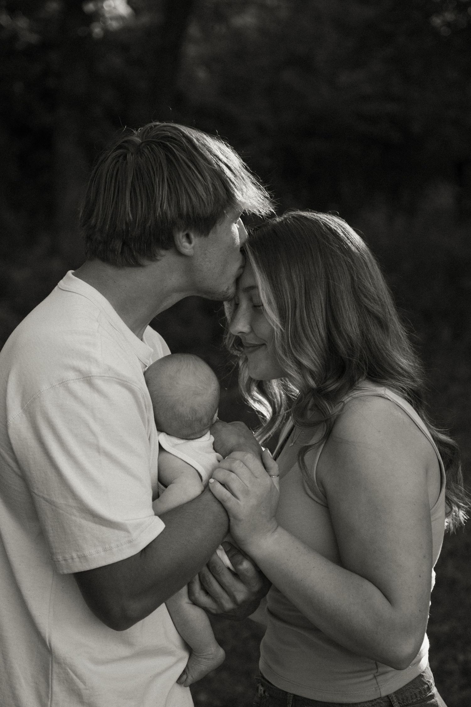 Black and white photo of new parents sharing a kiss while cradling their newborn baby.
