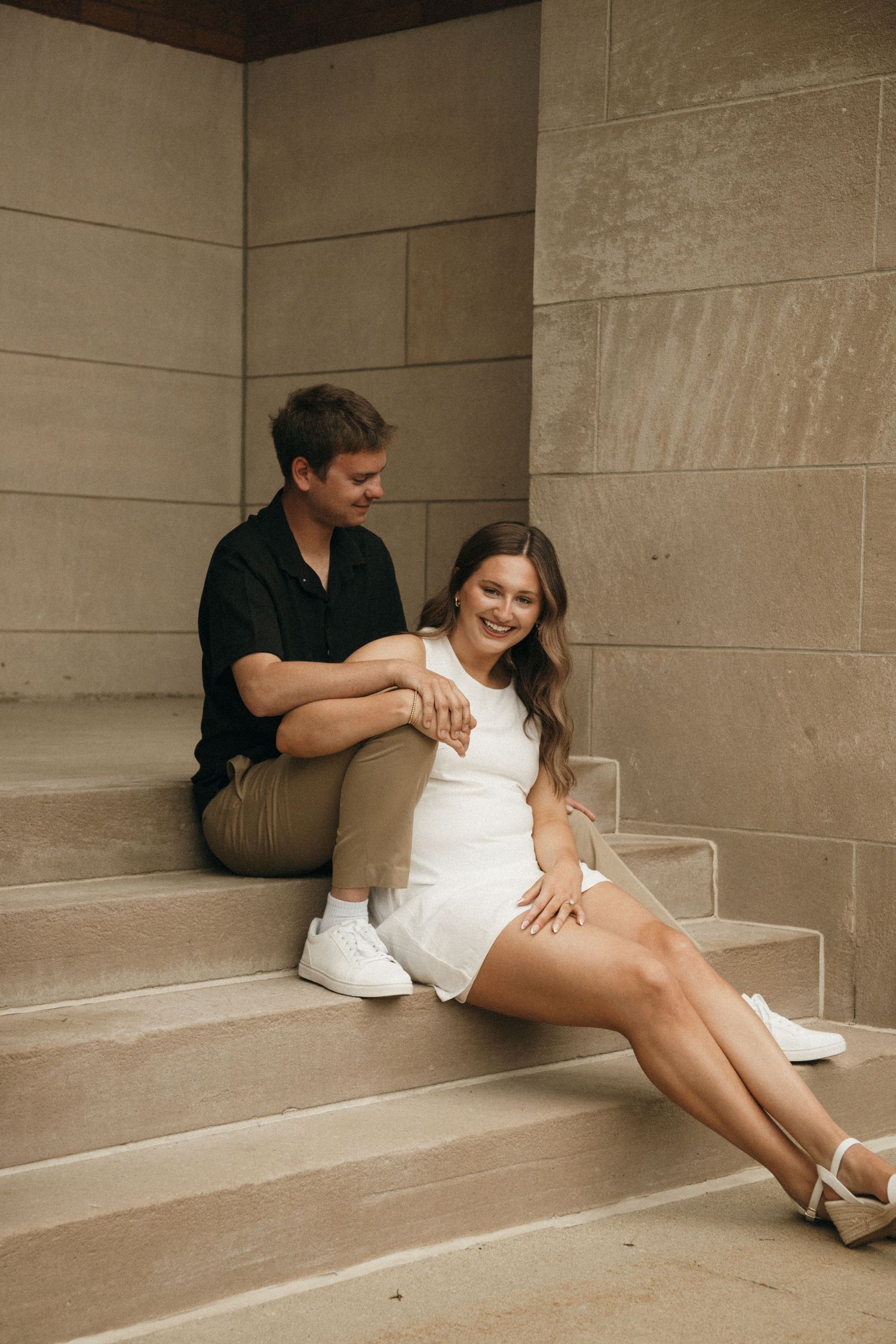 A couple sits together on stone steps, the woman in a white dress smiling as the man in black looks at her adoringly.