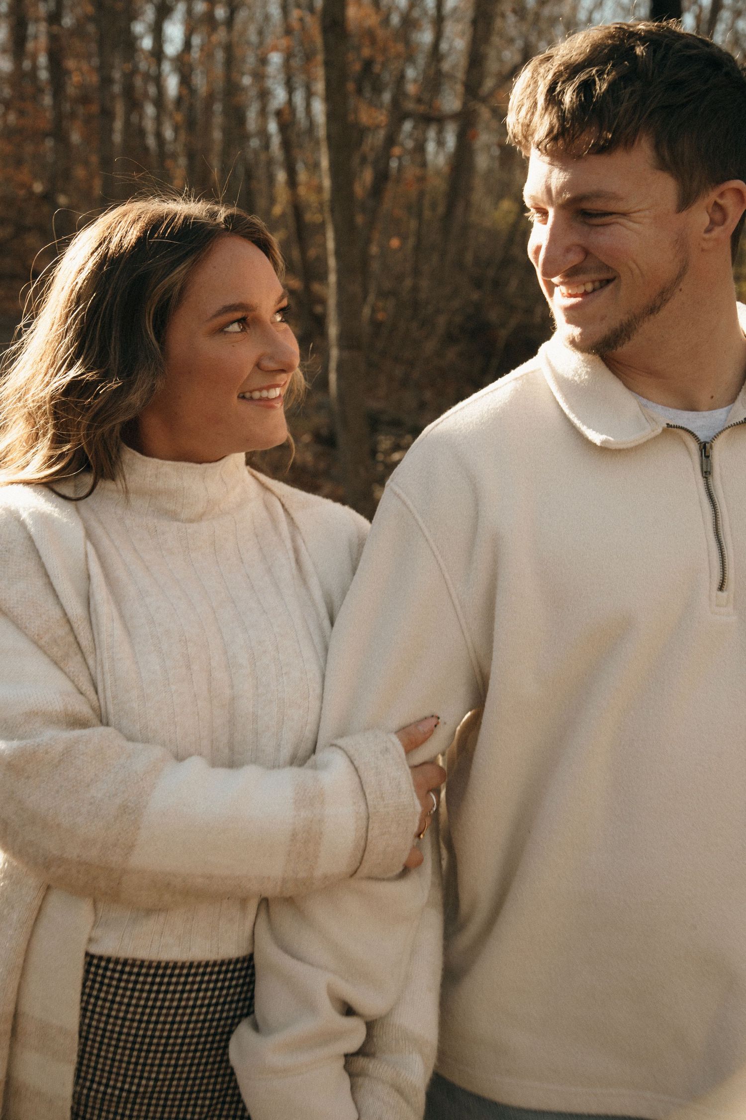 A candid moment of a couple in cream-colored sweaters sharing laughter in an autumn forest setting.
