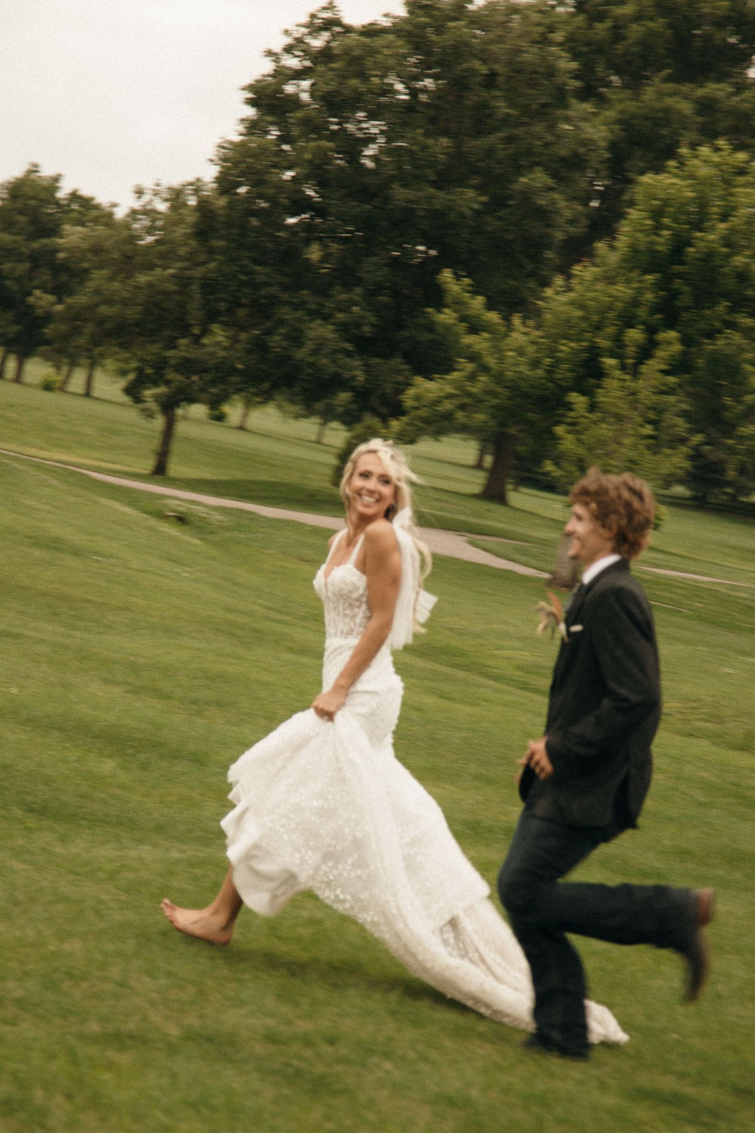 Joyful bride and groom running through lush green park on their wedding day.