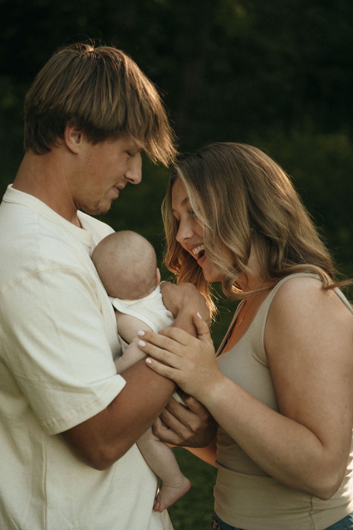 Young parents share a tender moment with their newborn baby outdoors in golden hour light.