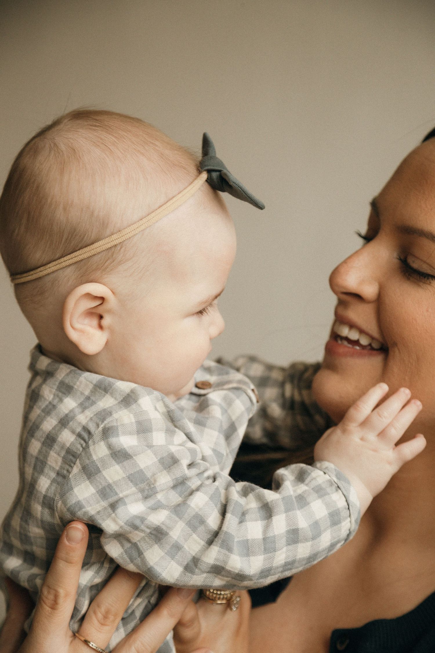 A smiling mother holds her baby in a gingham outfit with a bow headband, sharing a joyful moment.