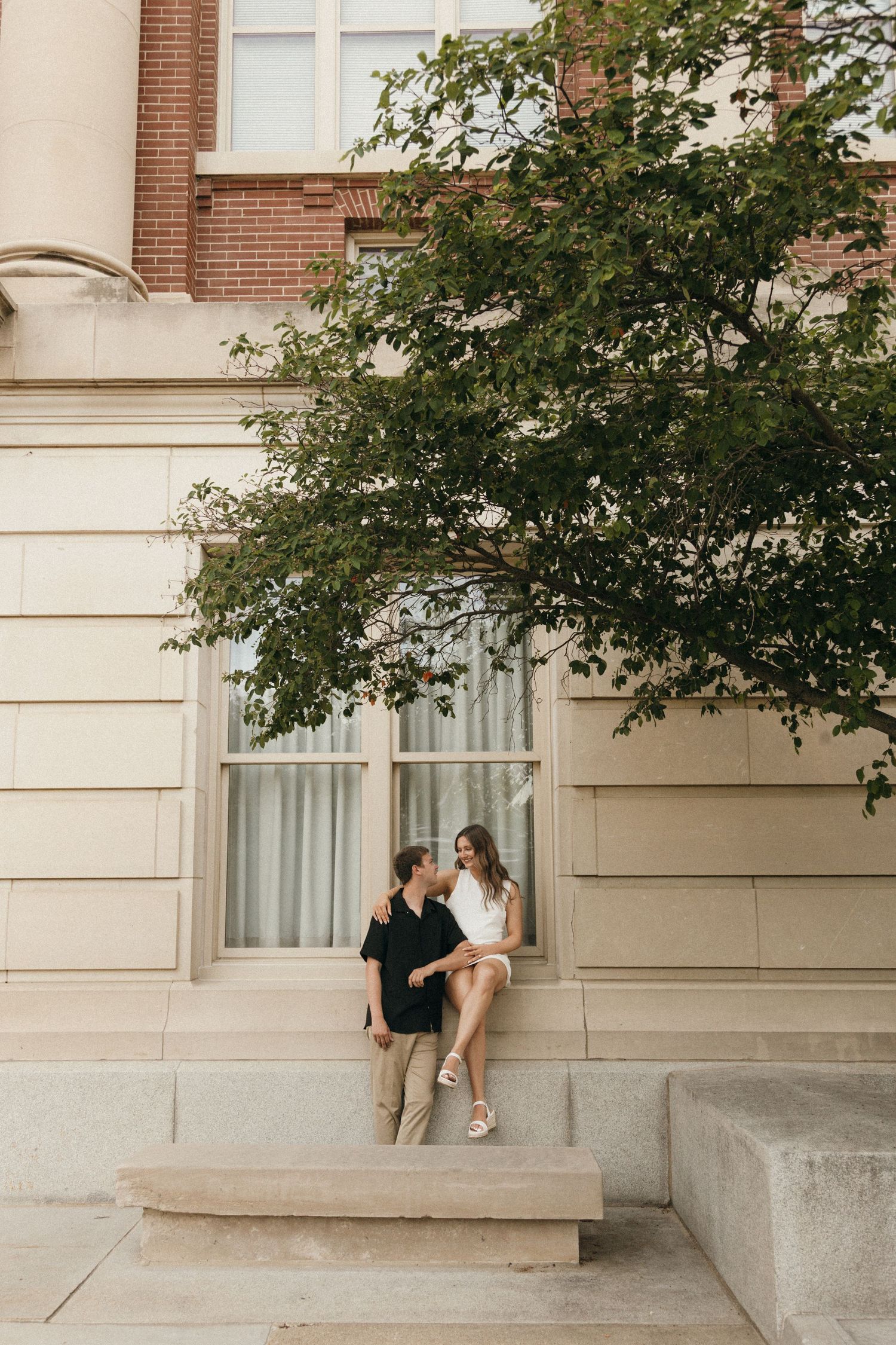 Couple sitting on stone steps outside a classic brick building, sharing a romantic moment under a lush green tree.