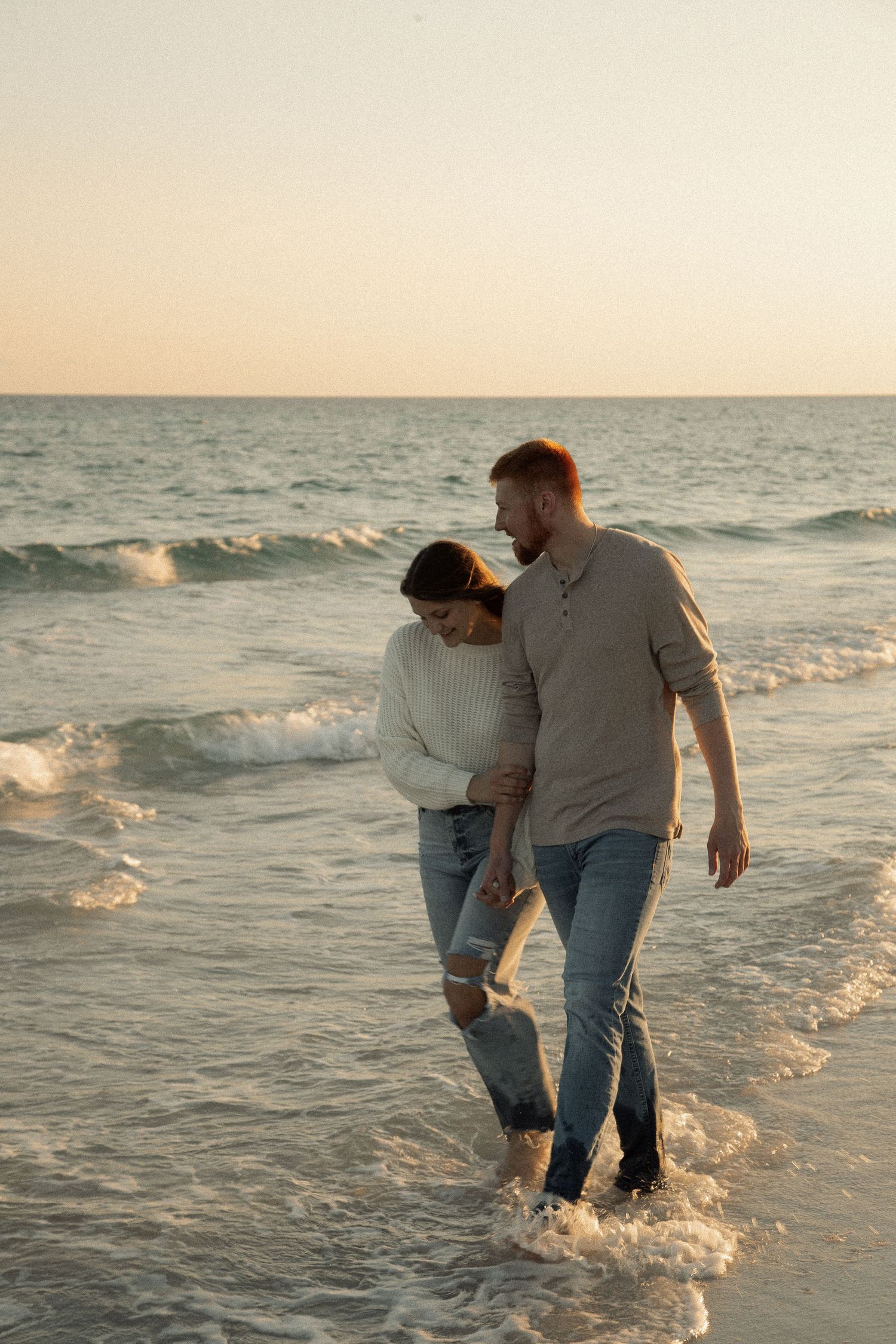 A couple walks together in the ocean waves at sunset along a peaceful beach.