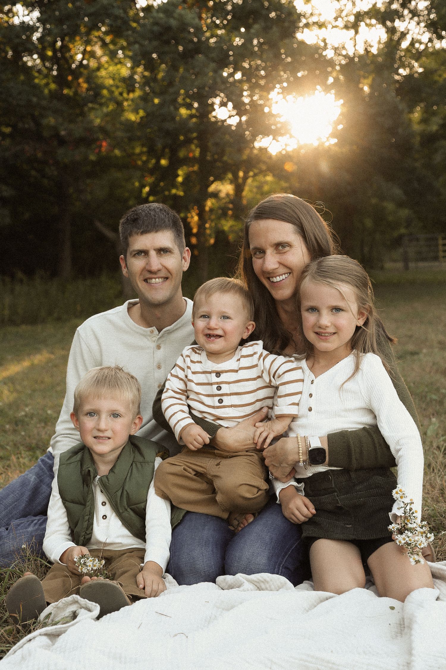 Family sitting together on white blanket during golden hour in park setting.