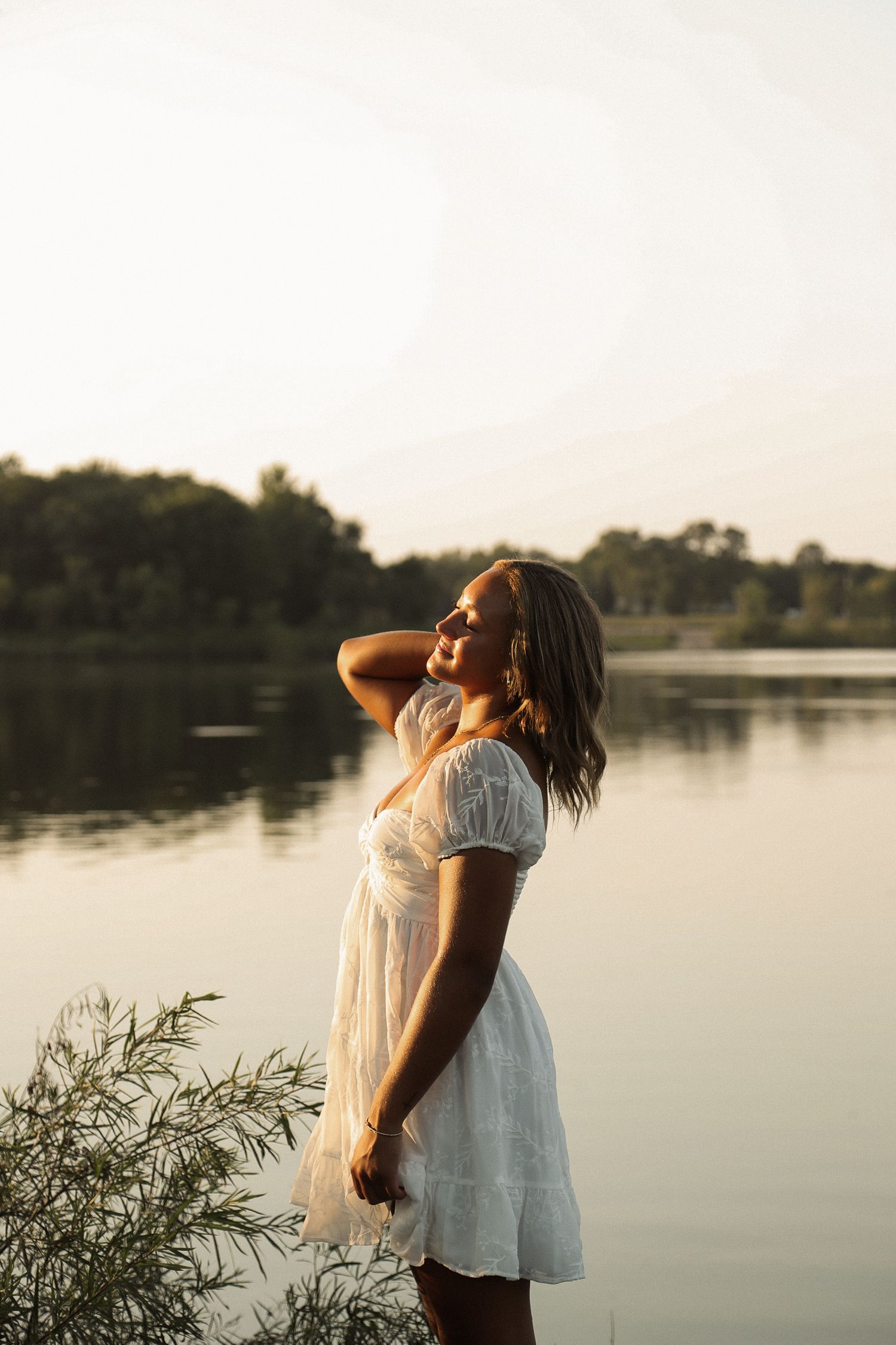 Person in white dress poses gracefully by a calm lake during golden hour.