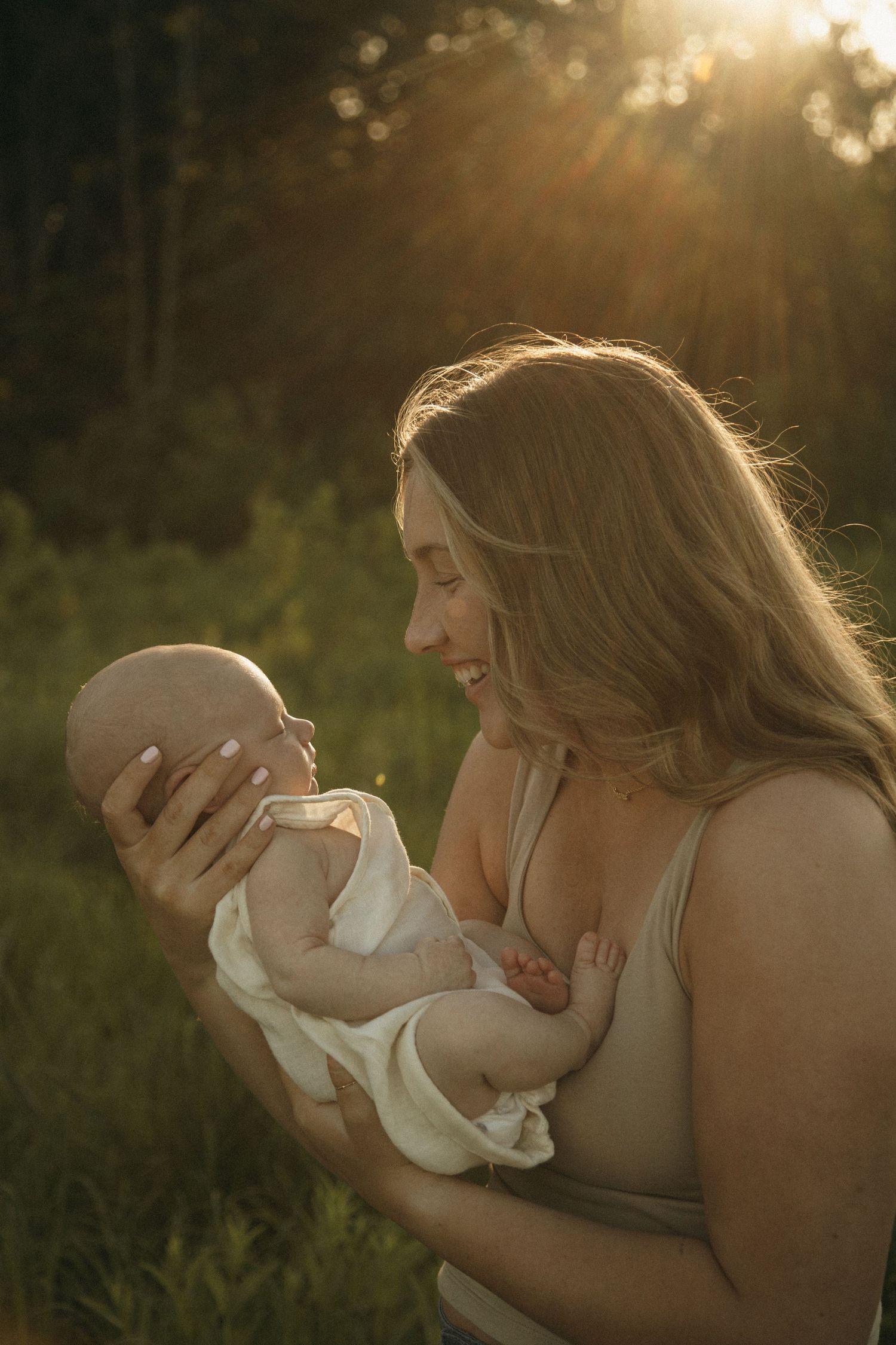 A smiling mother holds her newborn baby in golden sunlight outdoors, surrounded by lush green foliage.