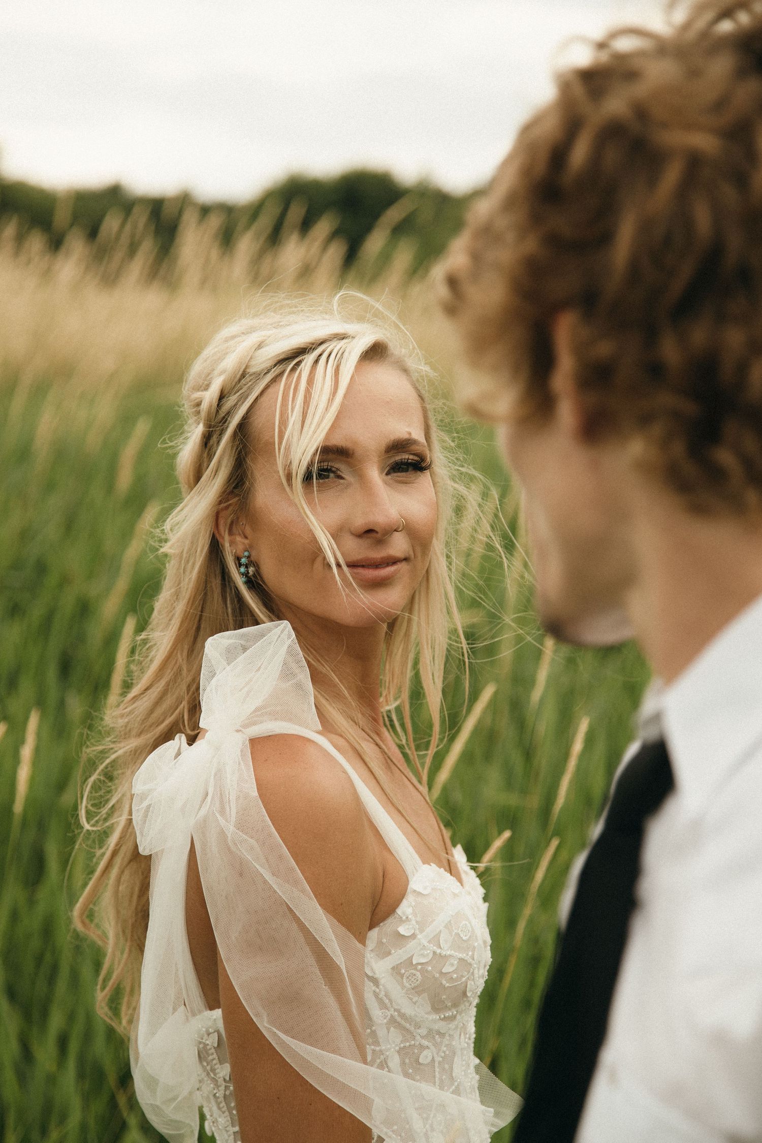 Blonde bride in lace wedding dress with flowing cape smiling at groom in tall grass field.