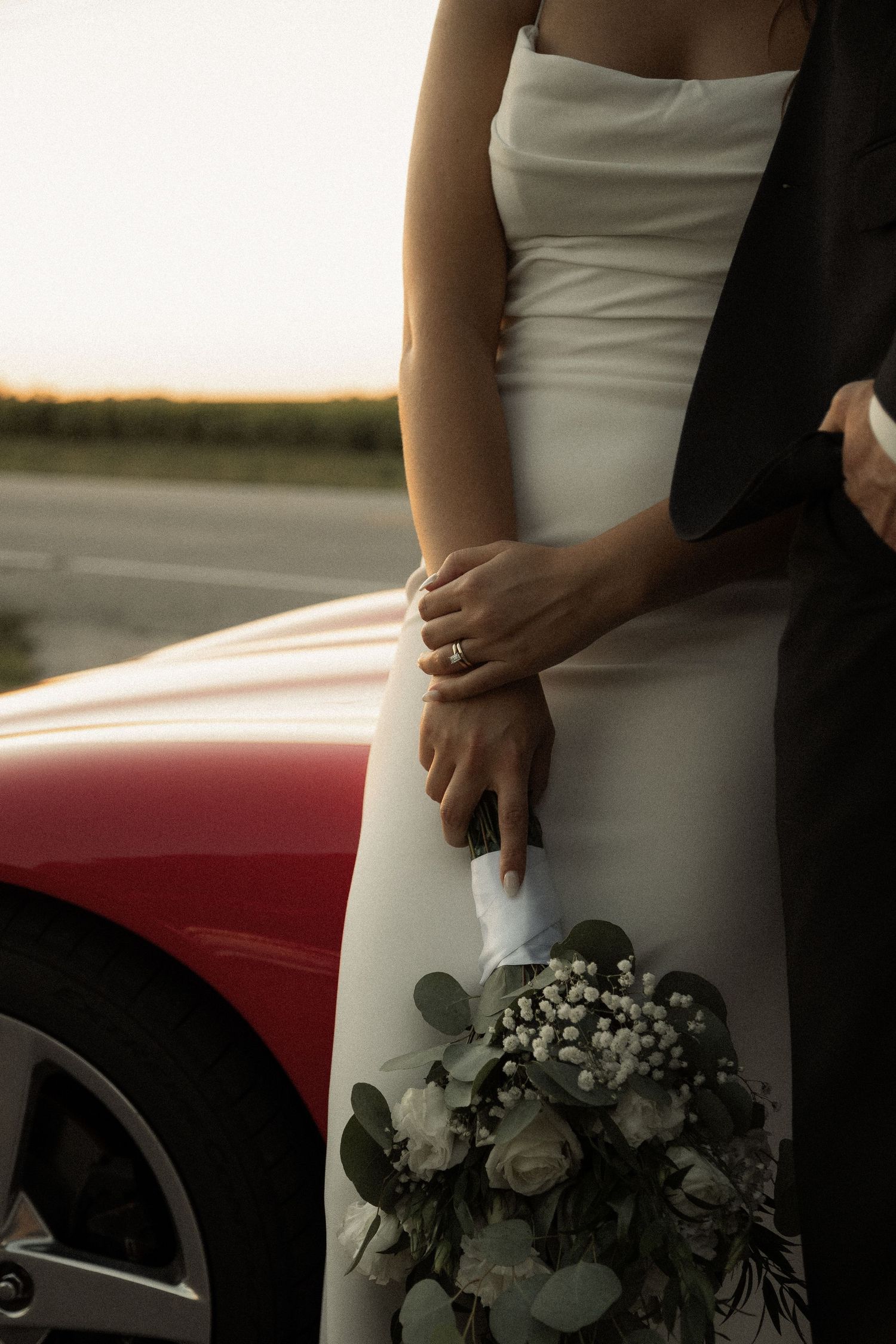 Detail shot of a white wedding dress and bouquet against a red car during sunset.