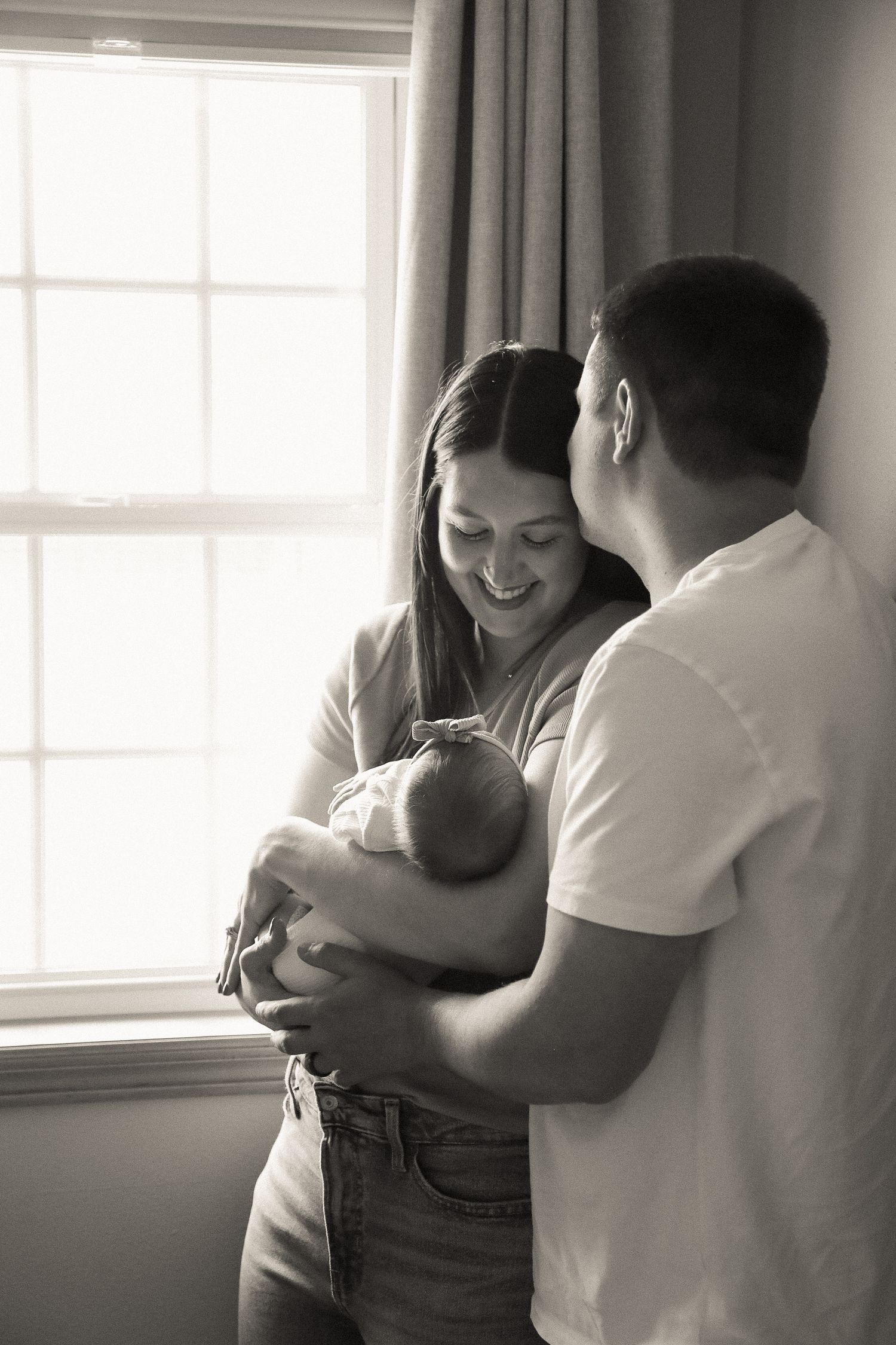 Parents share an intimate moment with their newborn by a window in soft natural light.