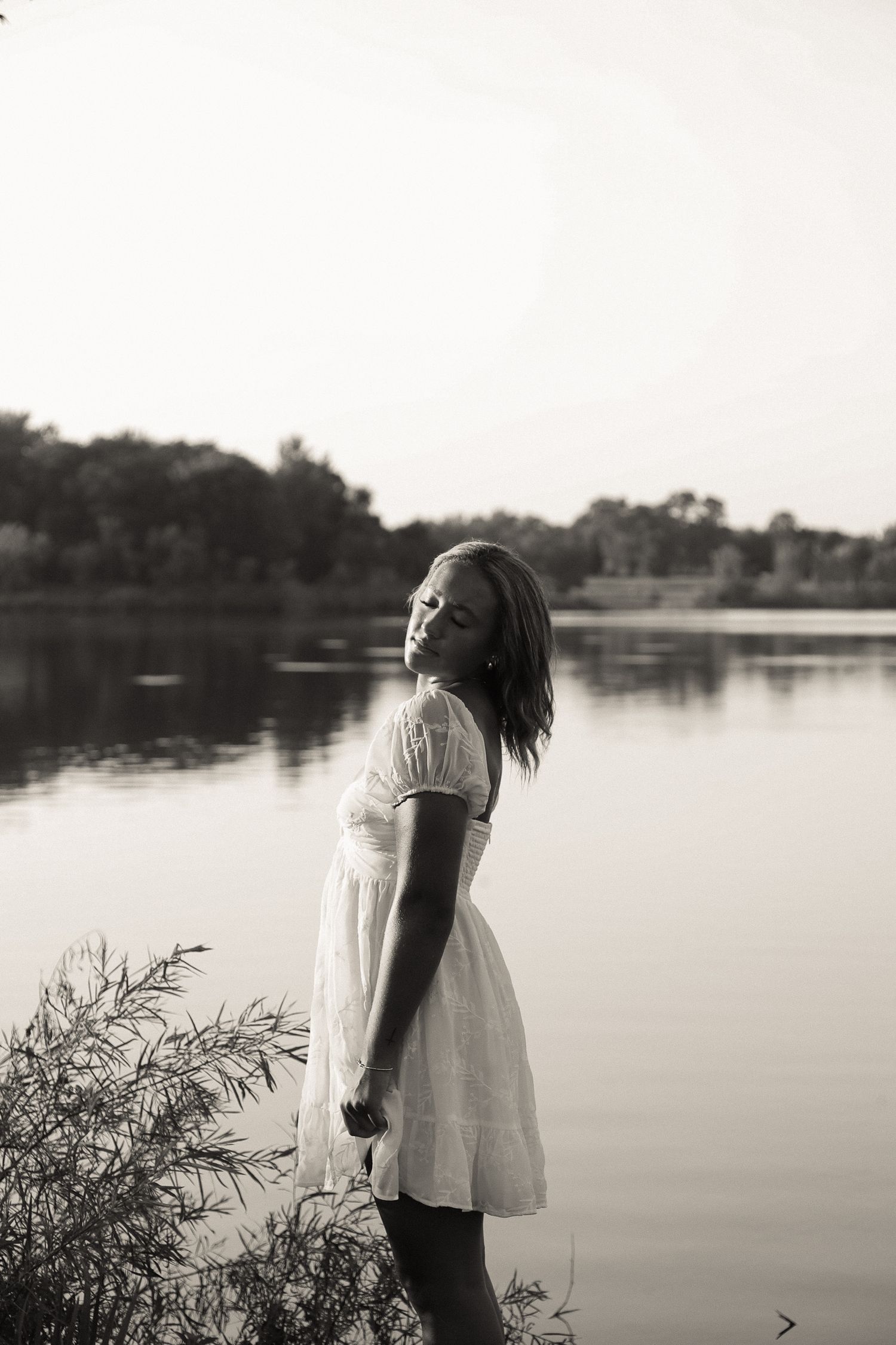 Dreamy black and white portrait by a serene lake with trees in the background.