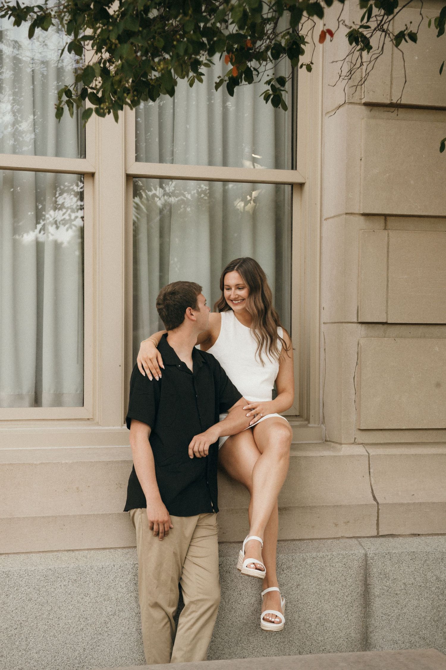 A couple shares a romantic moment outside a elegant building, woman in white dress seated on ledge, man in black shirt beside her.