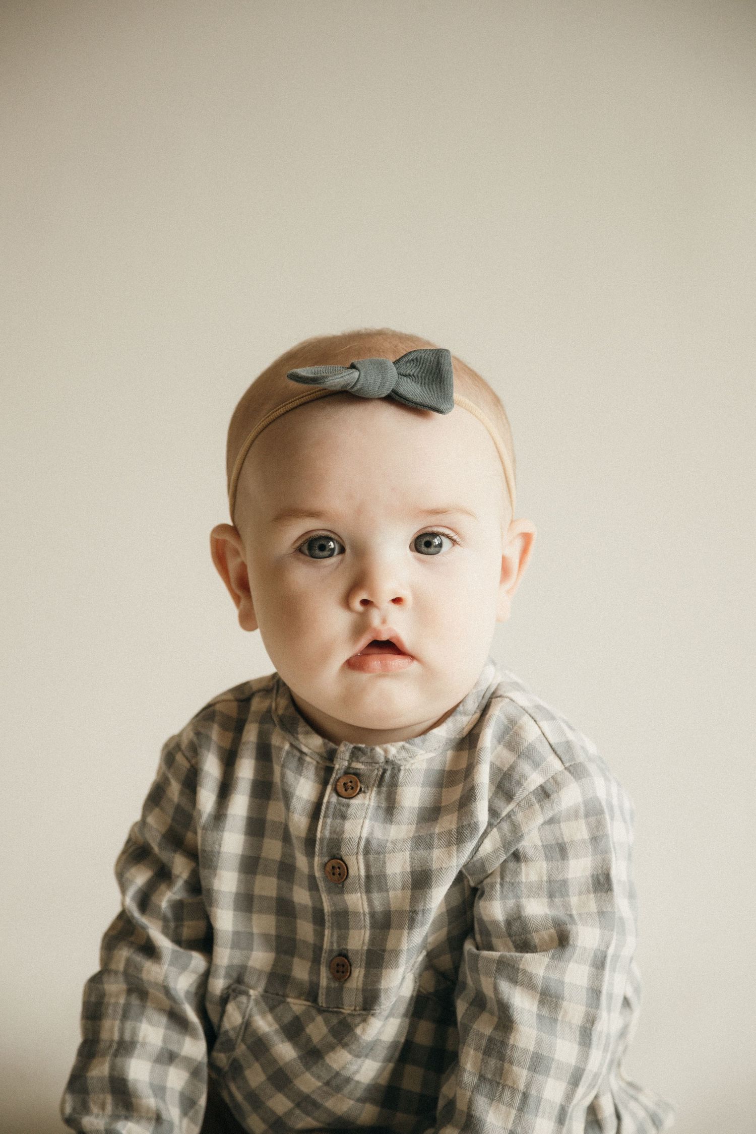 Adorable baby girl wearing a plaid outfit and gray bow headband, sitting against a neutral background.