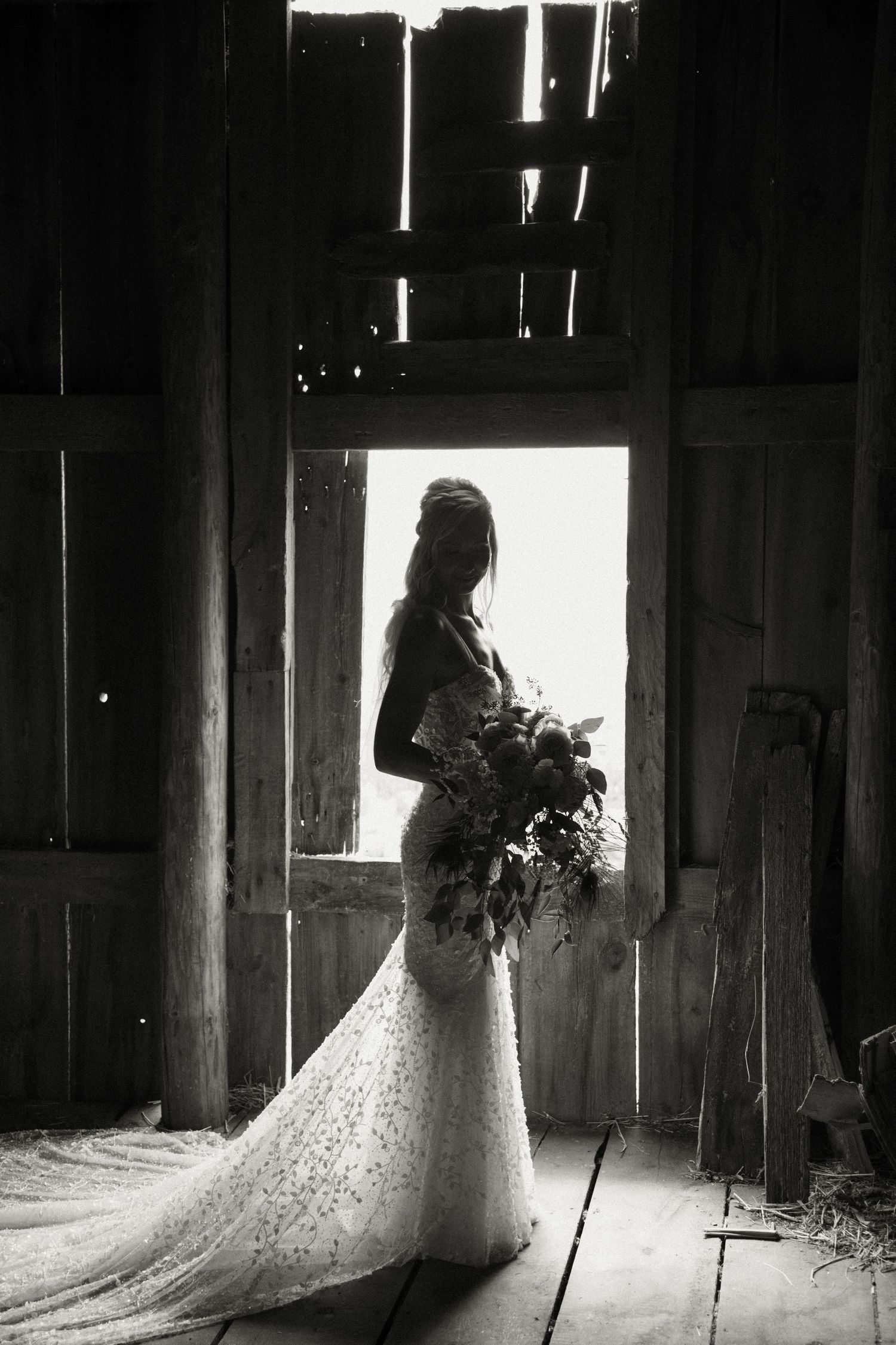 Bride in lace gown holding bouquet silhouetted in rustic barn doorway, black and white wedding photo.
