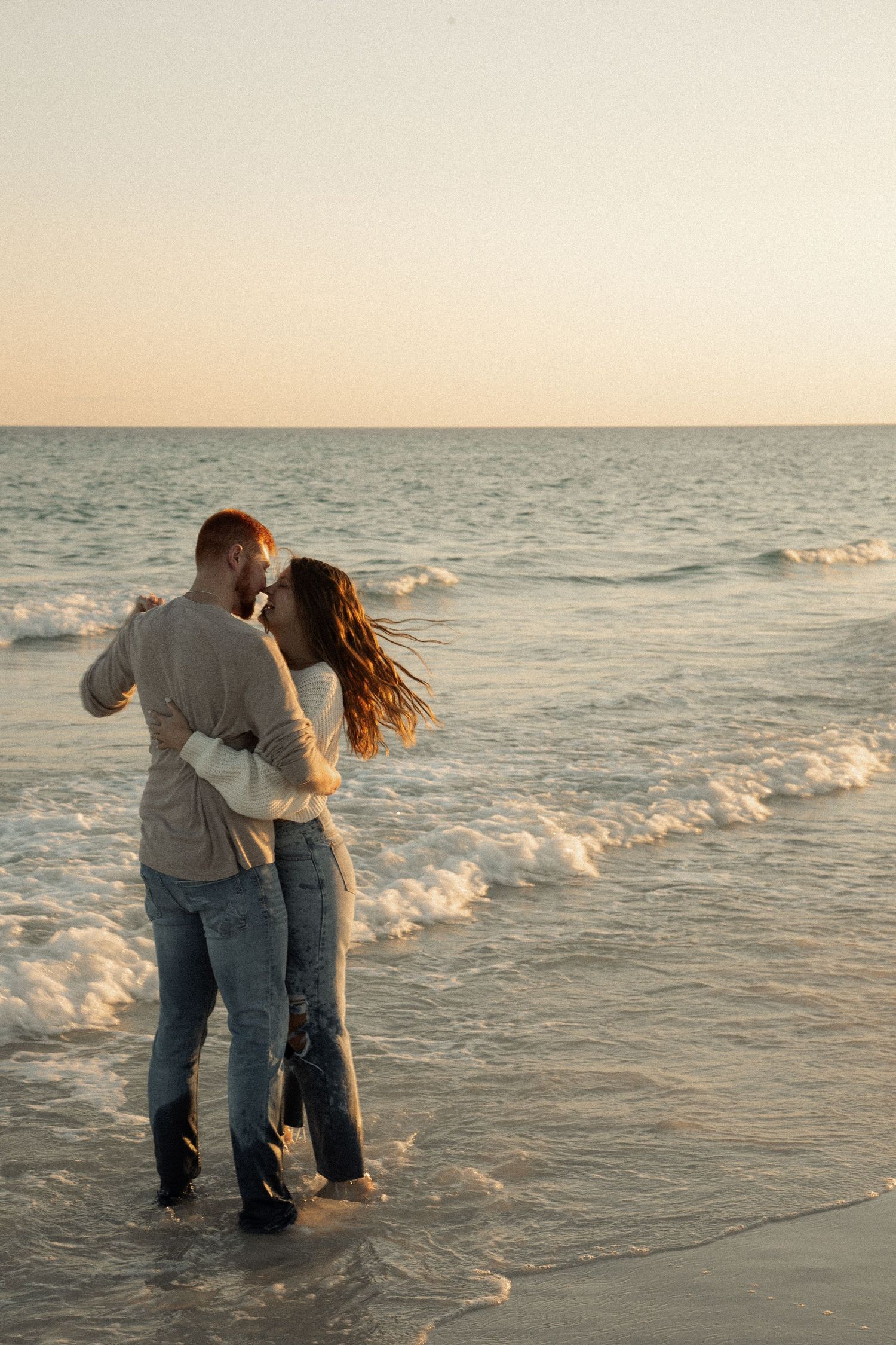 Couple embracing while standing in shallow ocean water at dusk.