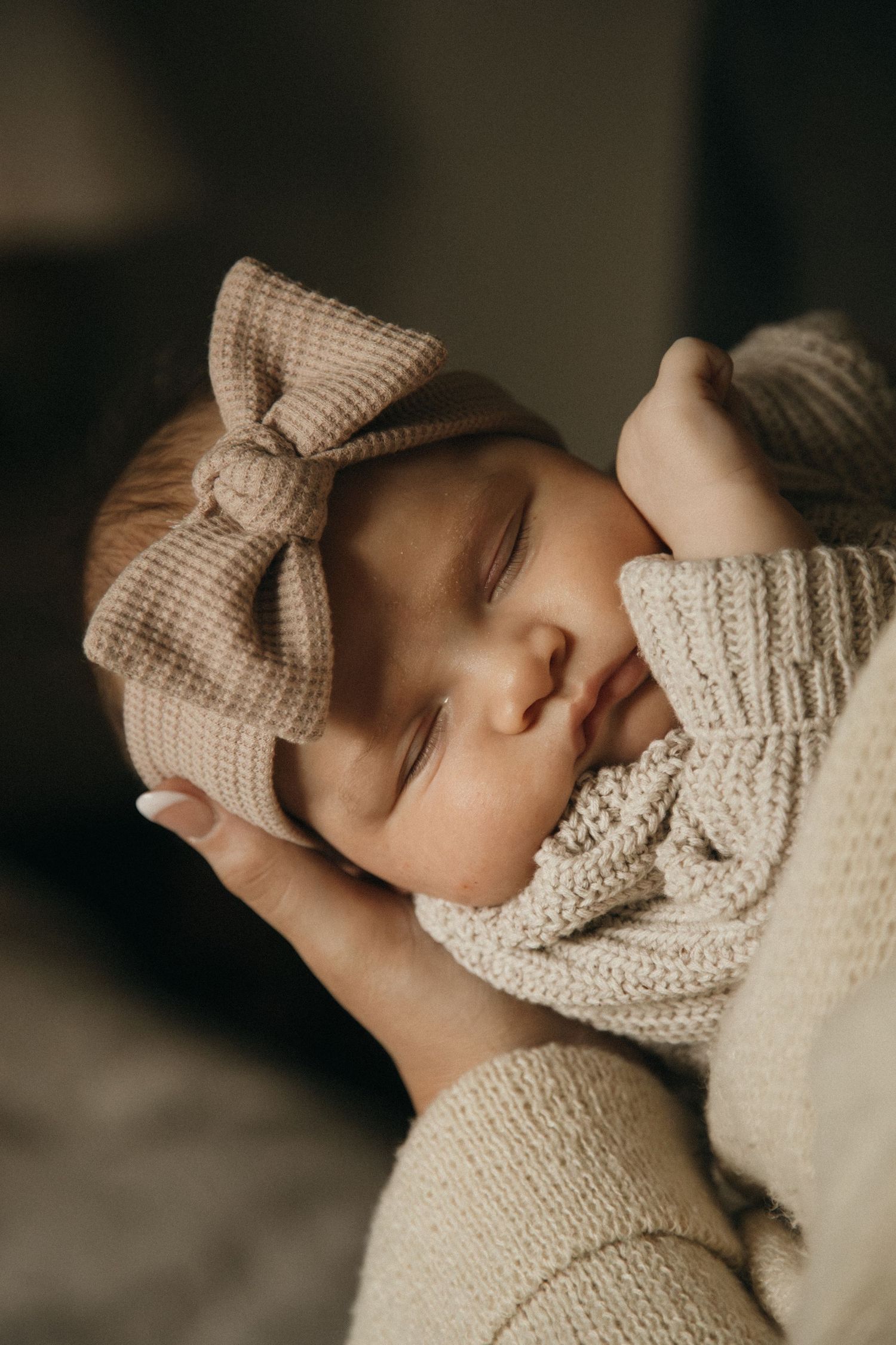 Close-up portrait of a sleeping newborn wearing a knitted bow headband and cream sweater.