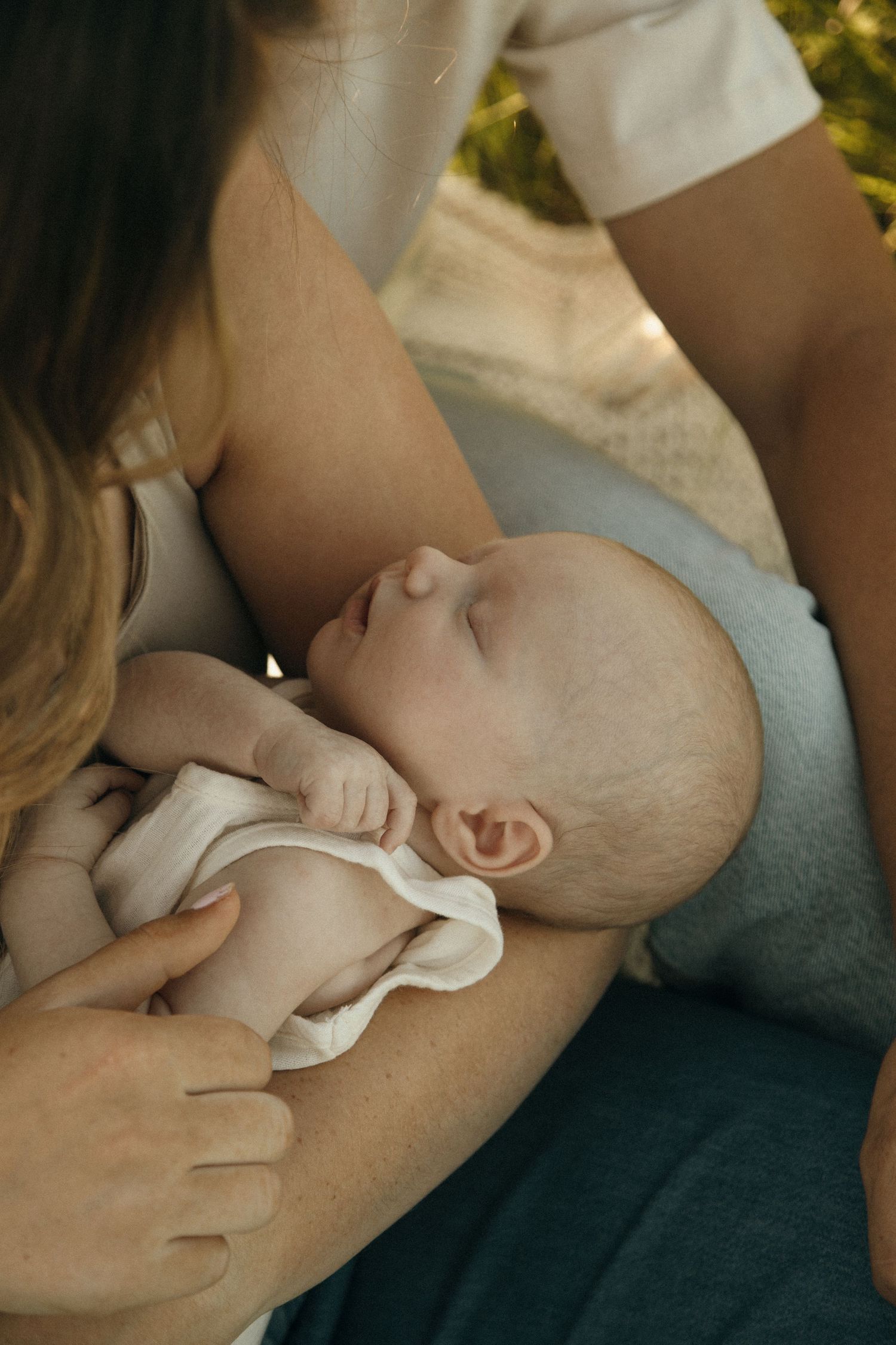 Peaceful newborn baby sleeping in parents arms outdoors, wearing white onesie, cradled lovingly.