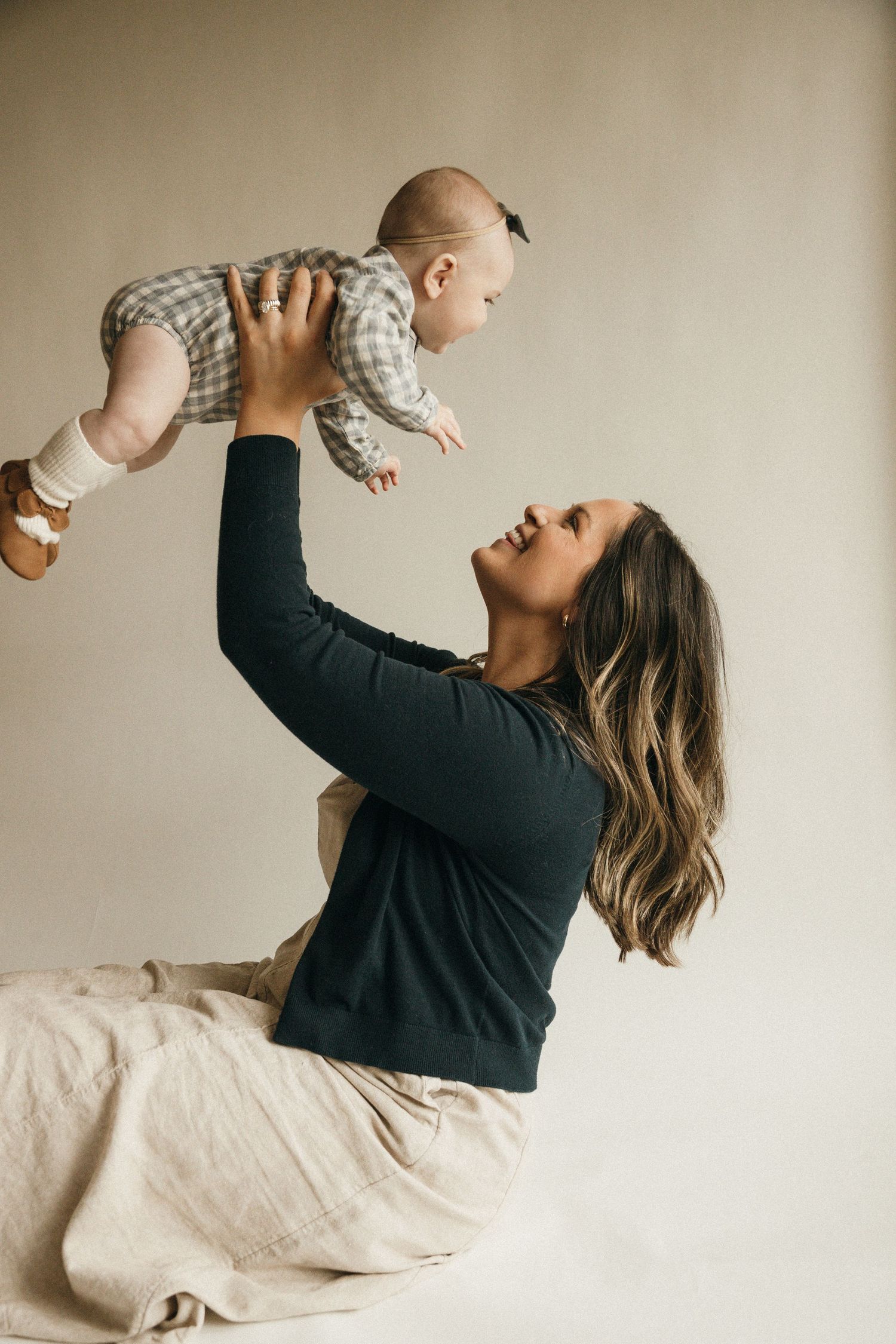 A smiling mother lifts her happy baby overhead while sitting on a beige blanket in a bright, neutral studio setting.