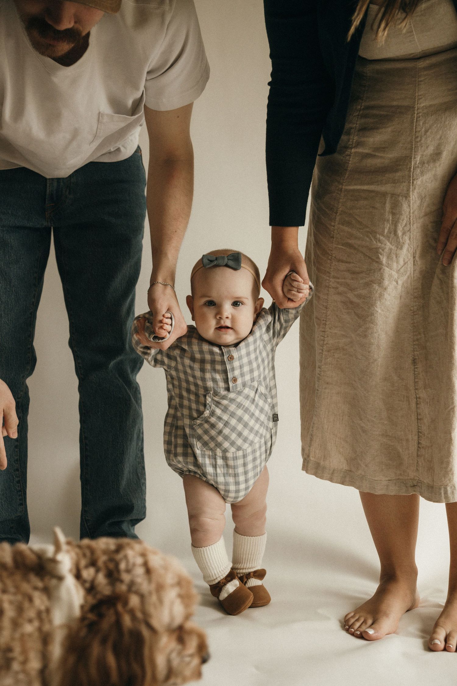 A baby in a gingham romper takes first steps while parents hold her hands in a bright white studio.