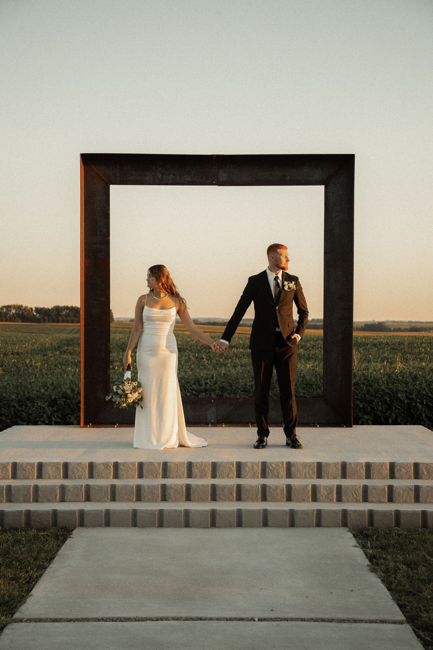 Wedding couple posing in modern black frame against sunset field backdrop.