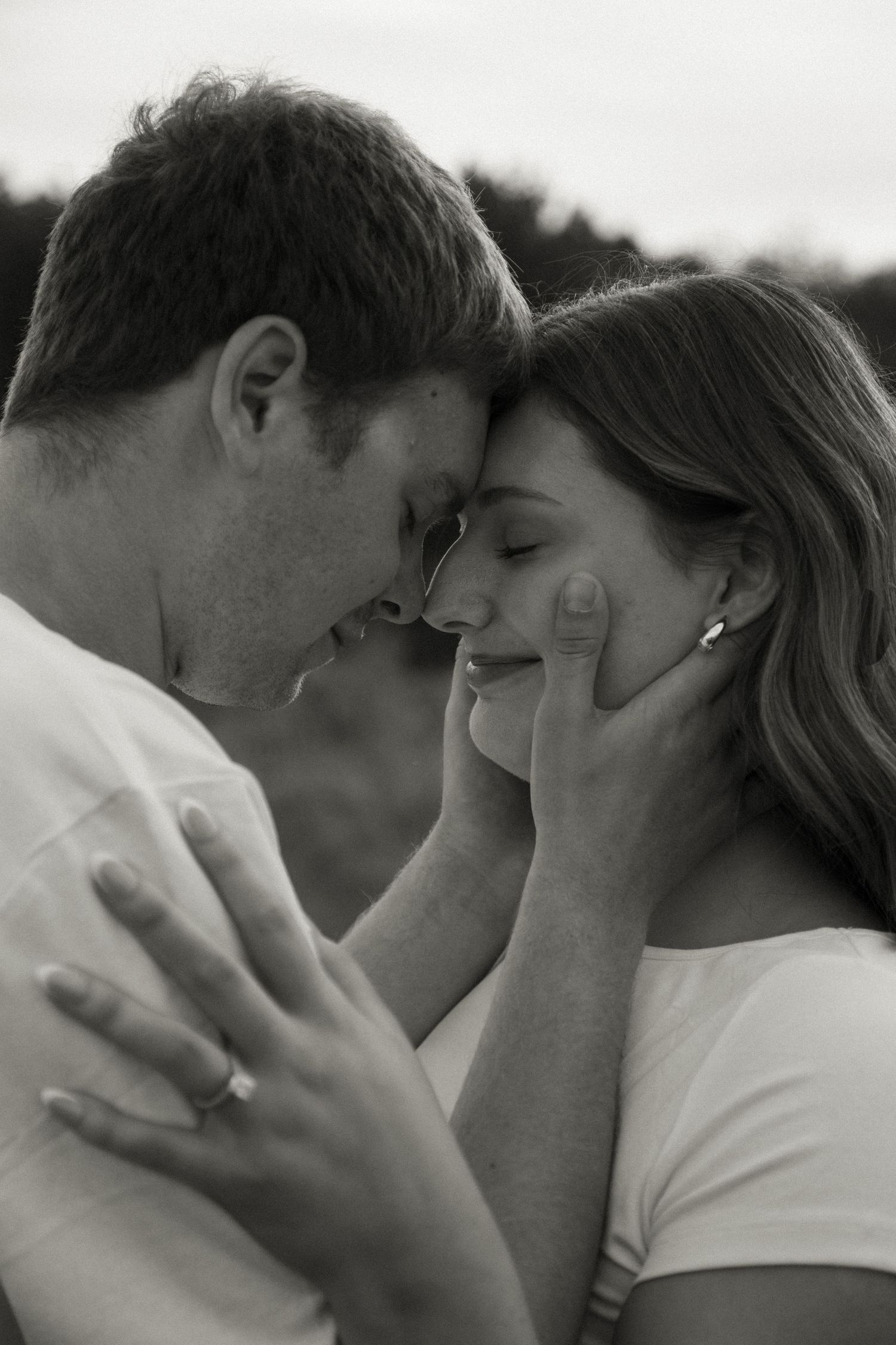 Black and white photo of a romantic couple touching foreheads tenderly outdoors.