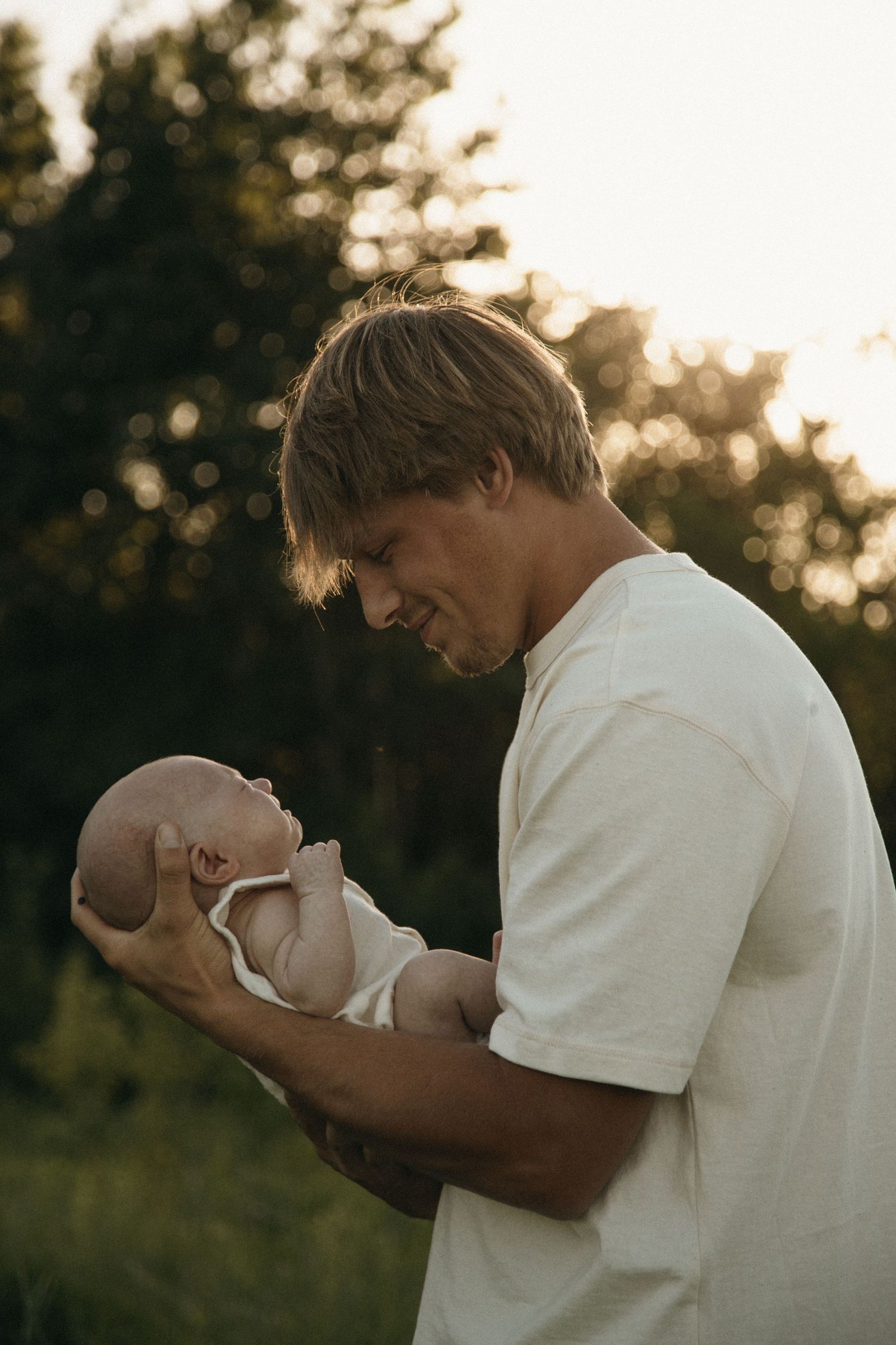 Father tenderly holding newborn baby in his hands outdoors in warm sunset light.