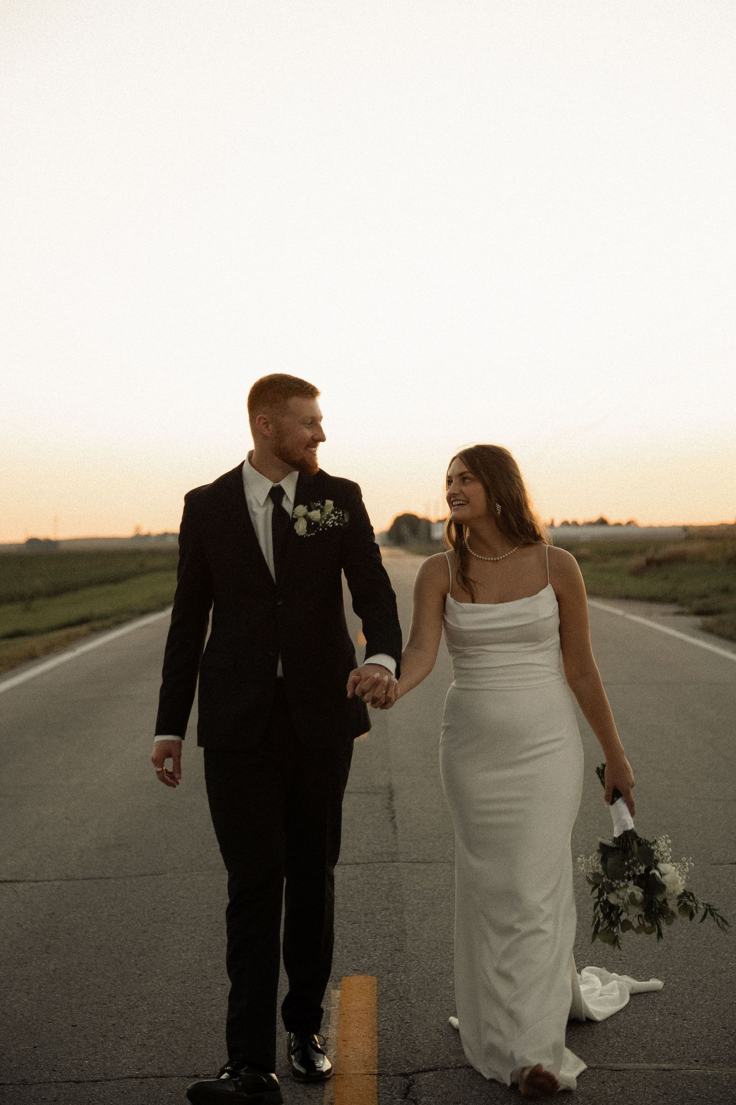 A couple in wedding attire walks along a rural road at sunset in a series of romantic photos.