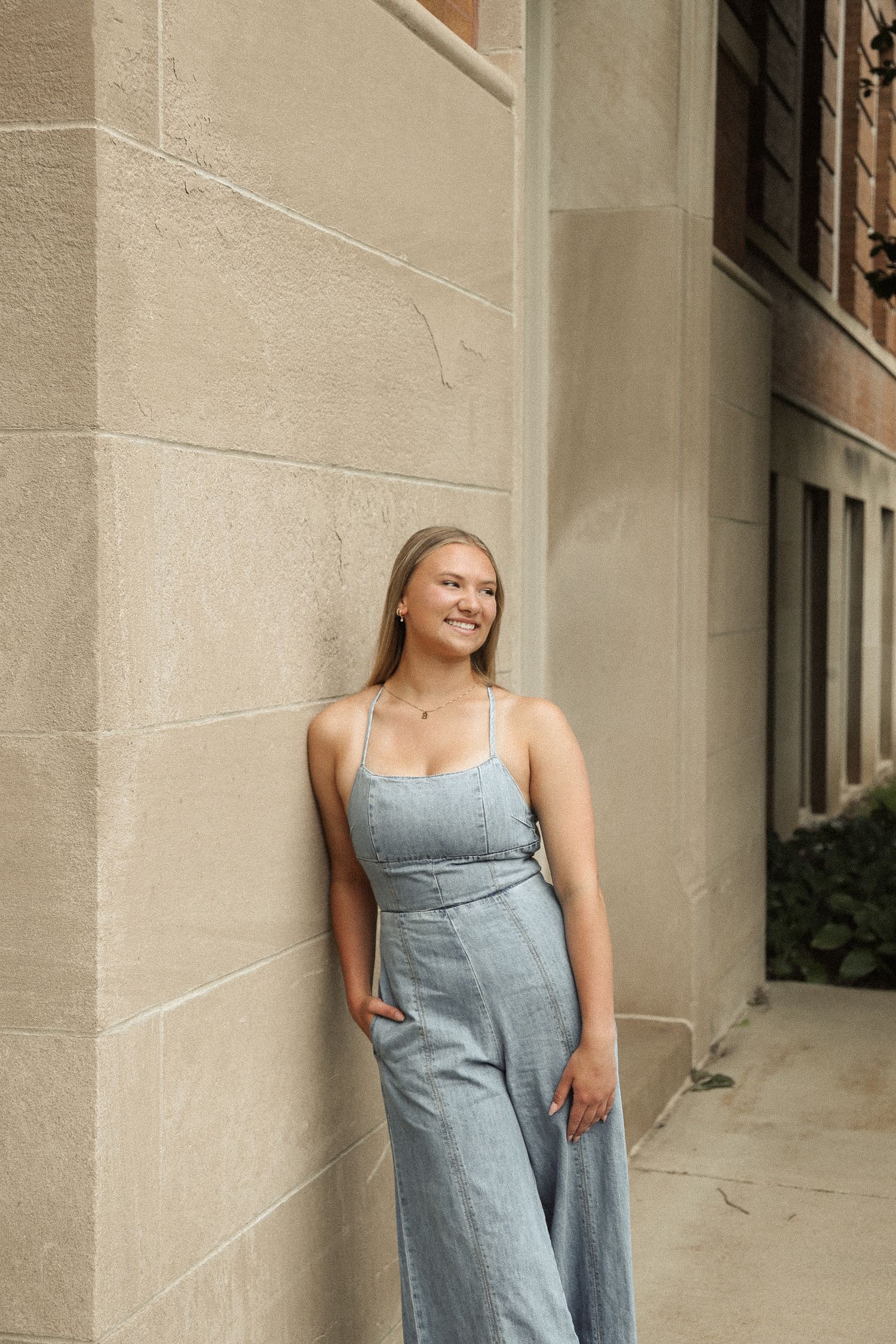 A woman in a denim dress poses playfully against a beige wall in natural light.