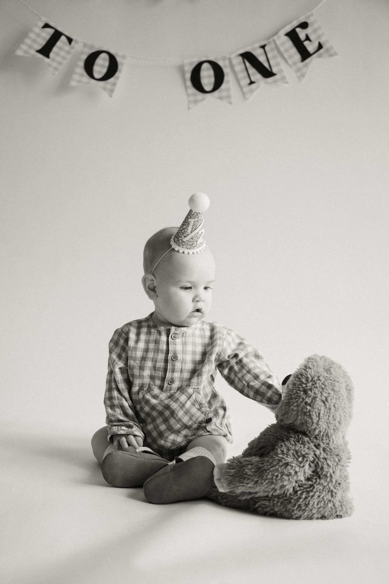 Black and white photo of a baby in a party hat sitting with a teddy bear under a 'To One' birthday banner.
