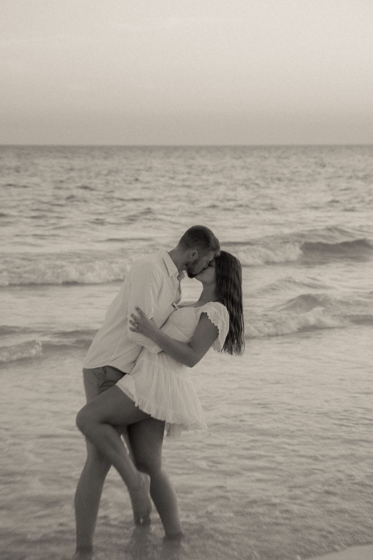 A romantic couple embraces while standing in ocean waves at sunset, creating a dreamy black and white scene.