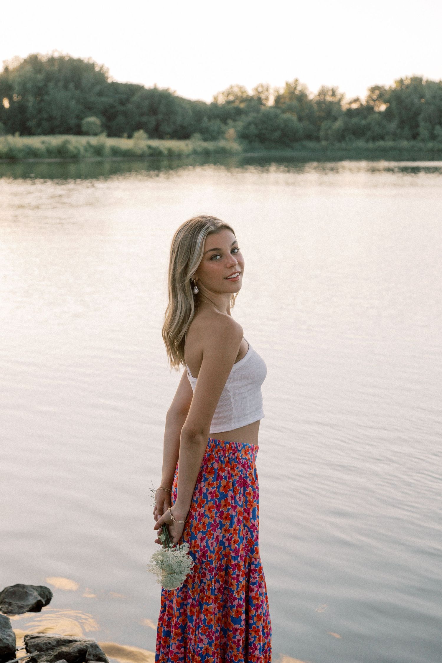 Person in white top and floral skirt poses by a serene lake at sunset.