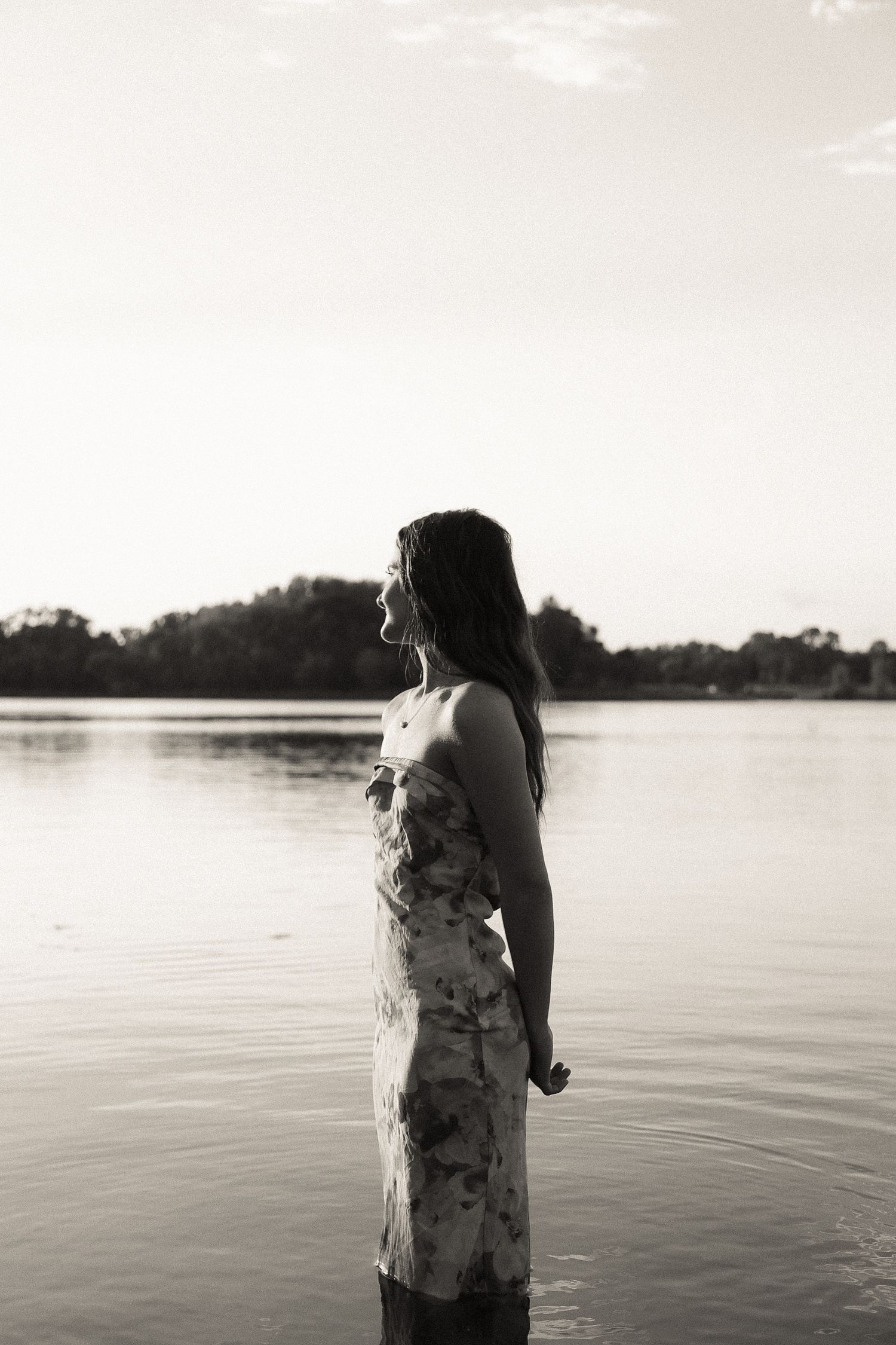 A silhouette stands by a serene lake at sunset in a flowing dress.