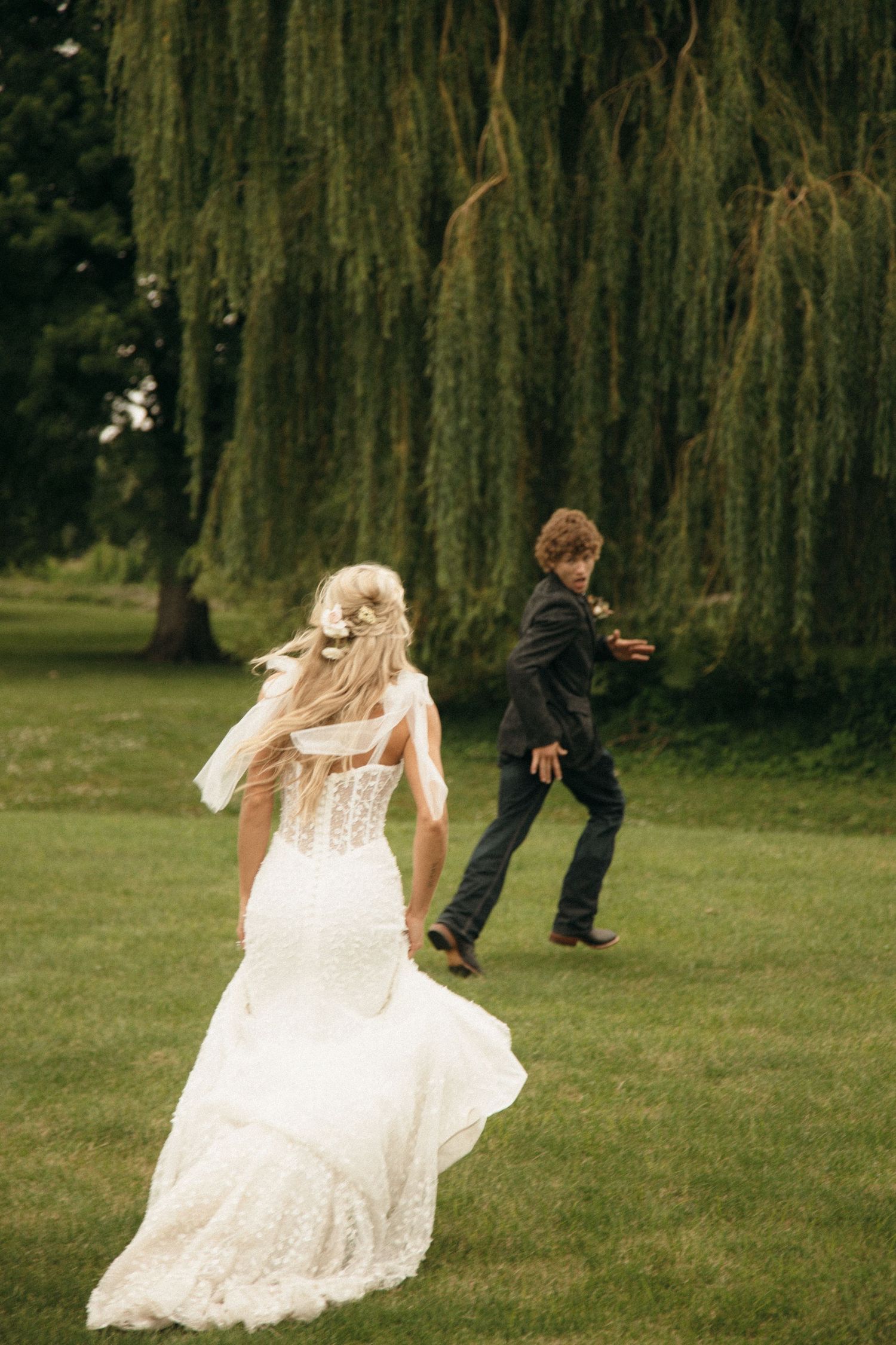Bride in lace gown running toward groom under weeping willow tree on green lawn.