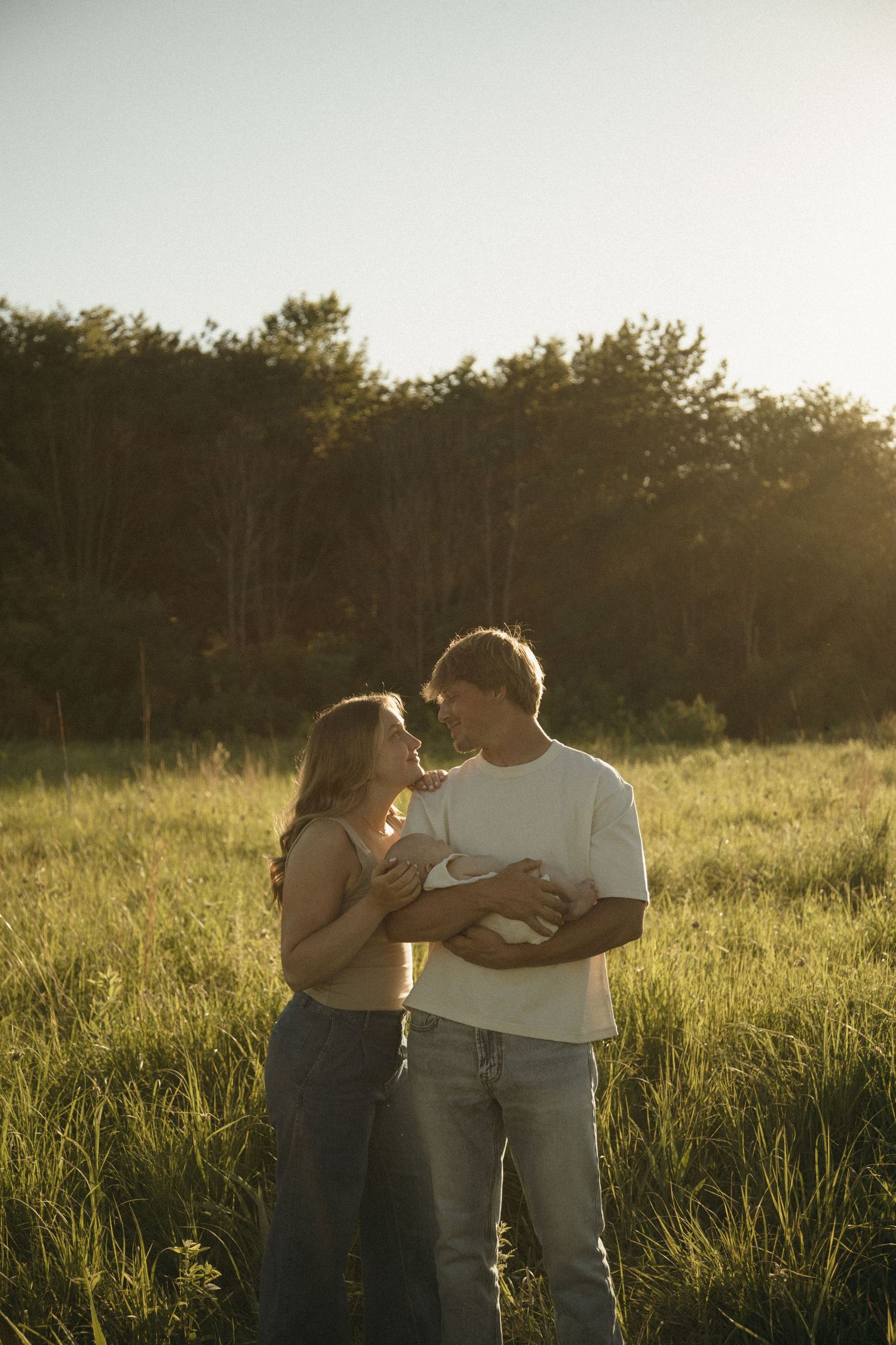 Couple standing in sunlit green field holding newborn baby, sharing tender moment with forest background.