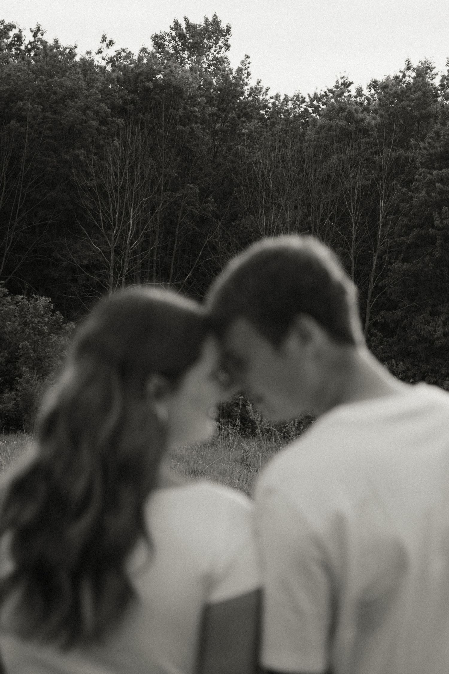 Black and white photo of a couple sharing an intimate moment, nearly kissing in front of a lush forest backdrop.