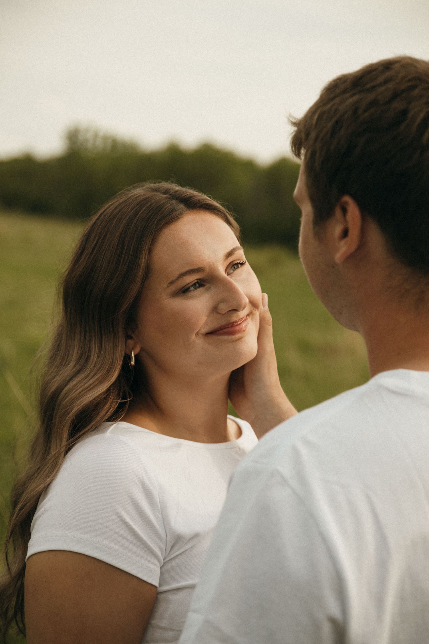 Young woman smiling warmly as her partner gently touches her face in a green field.