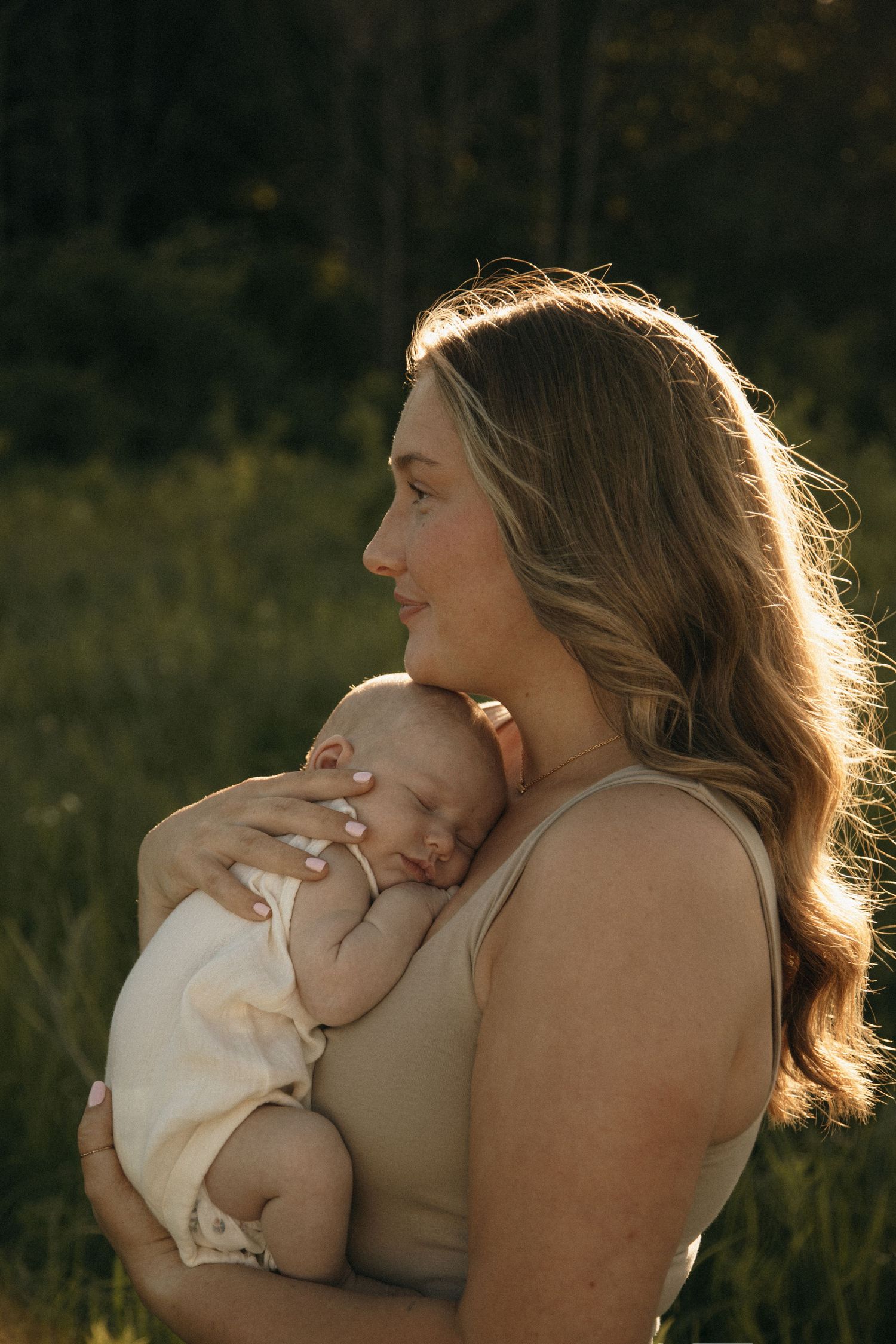 Mother holding sleeping newborn baby against her chest in golden sunset light outdoors.