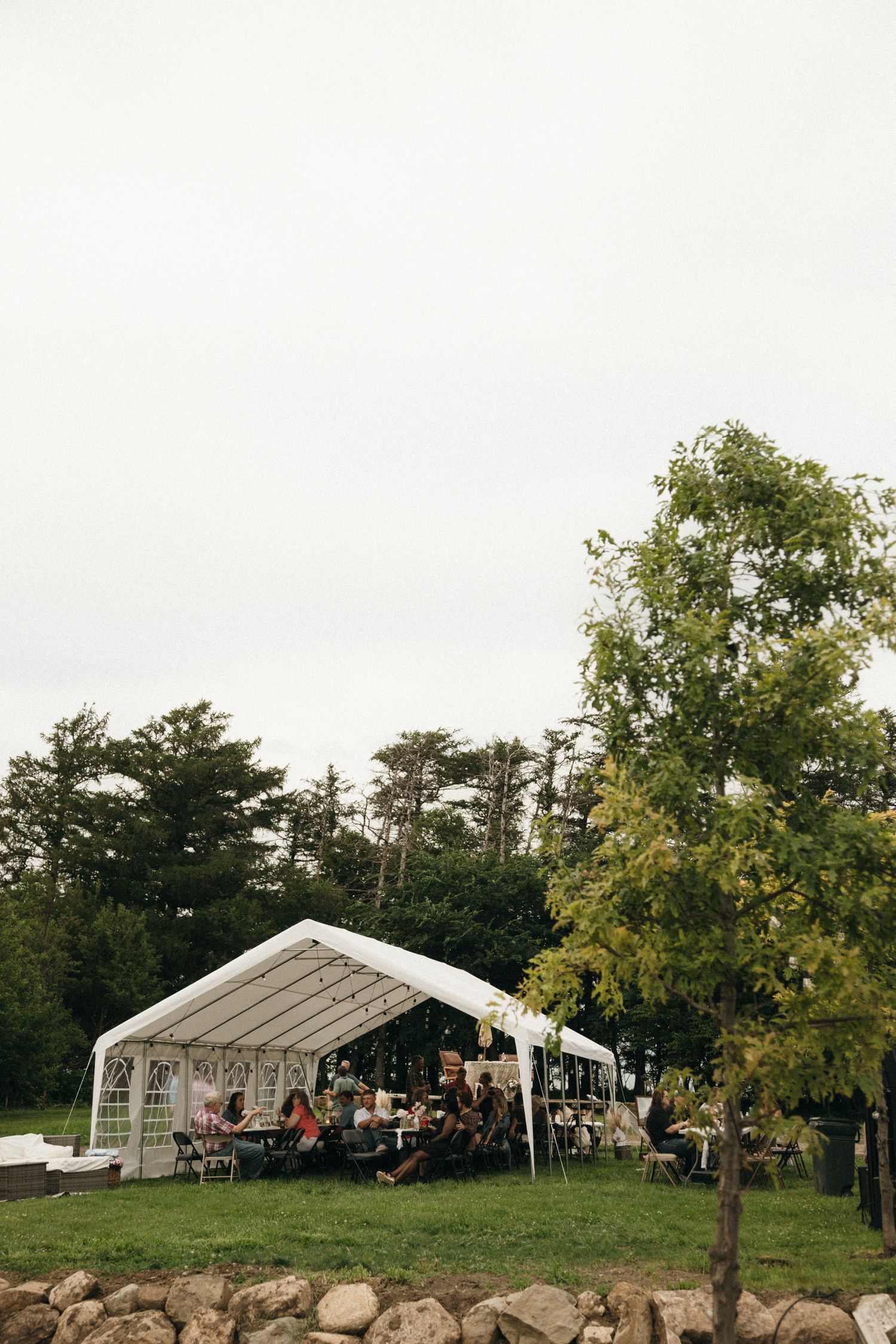Outdoor wedding reception under a white tent surrounded by lush green trees on an overcast day.