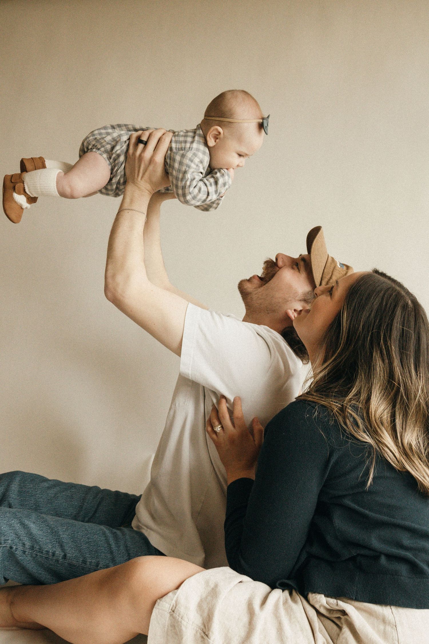 Happy parents sitting together, father lifting smiling baby in plaid outfit while mother leans in for a kiss.