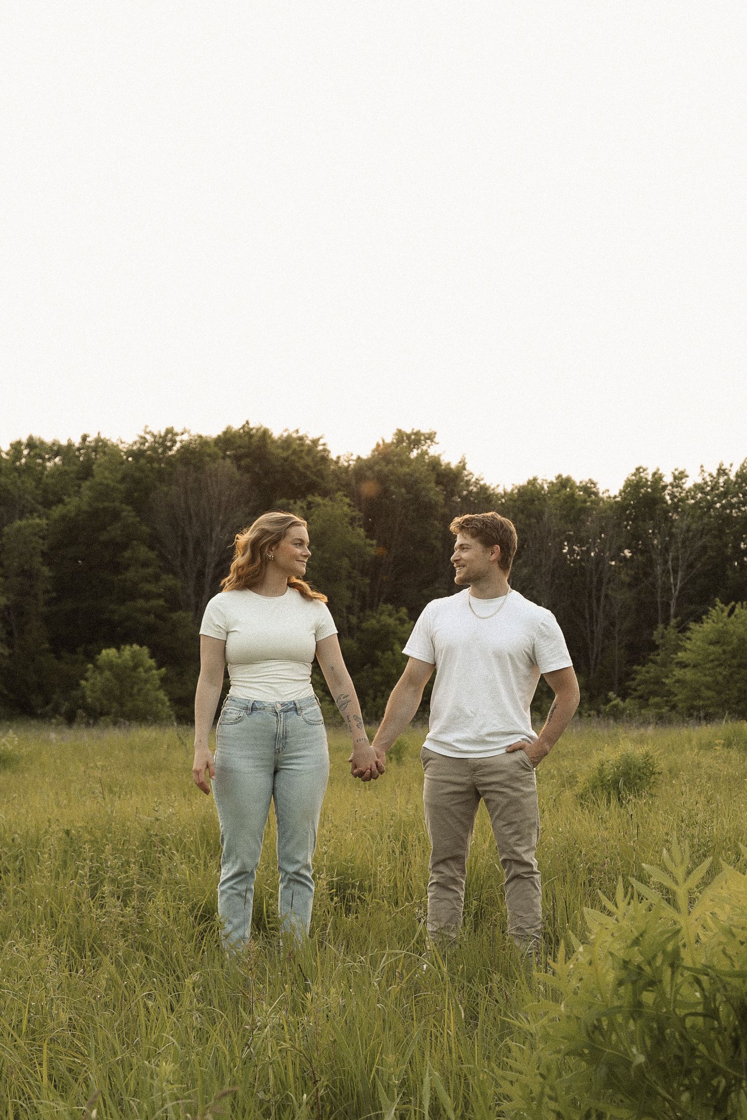 Two people holding hands while standing in a tall grass field with trees in the distance.