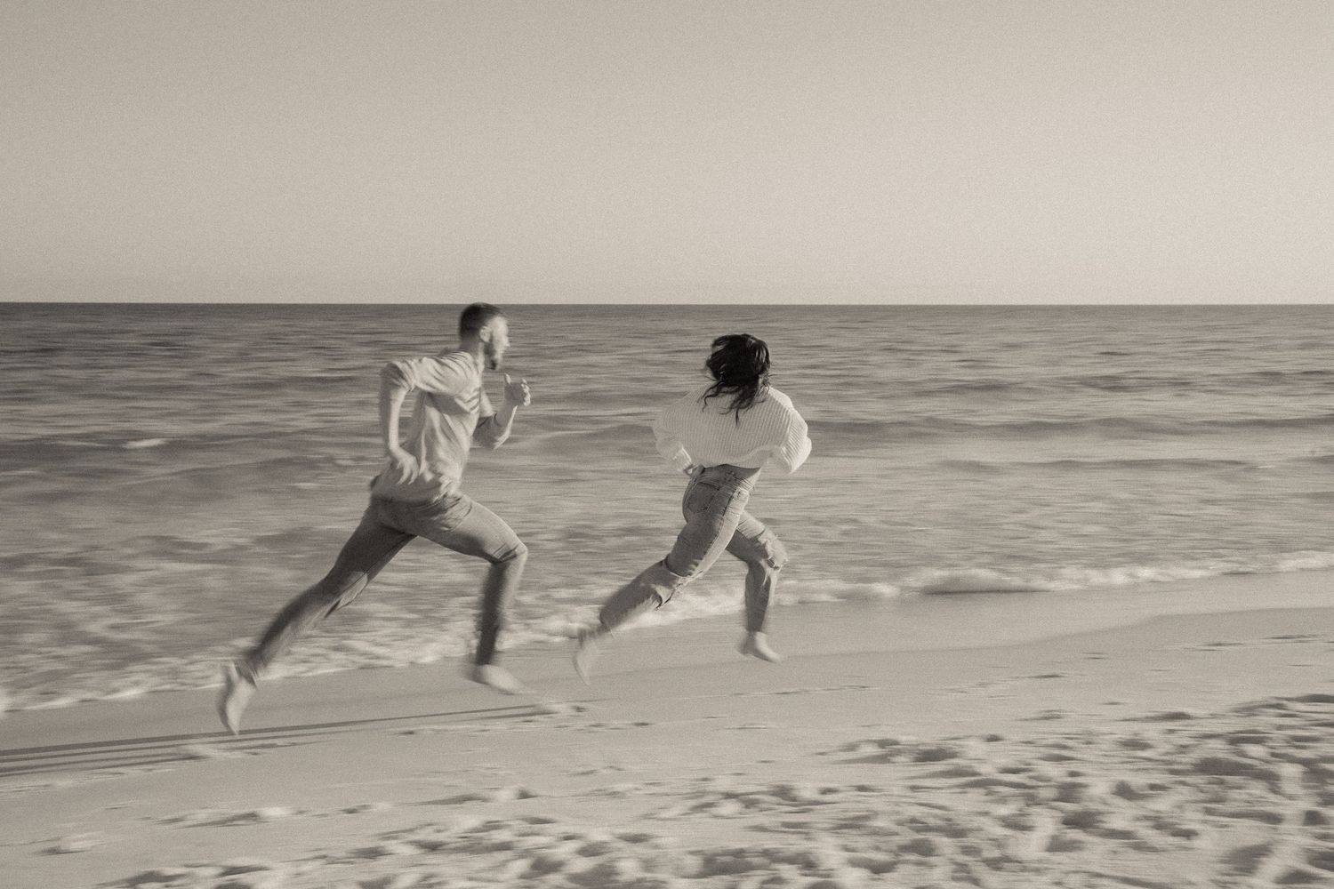 People running joyfully along a beach at sunset in black and white.