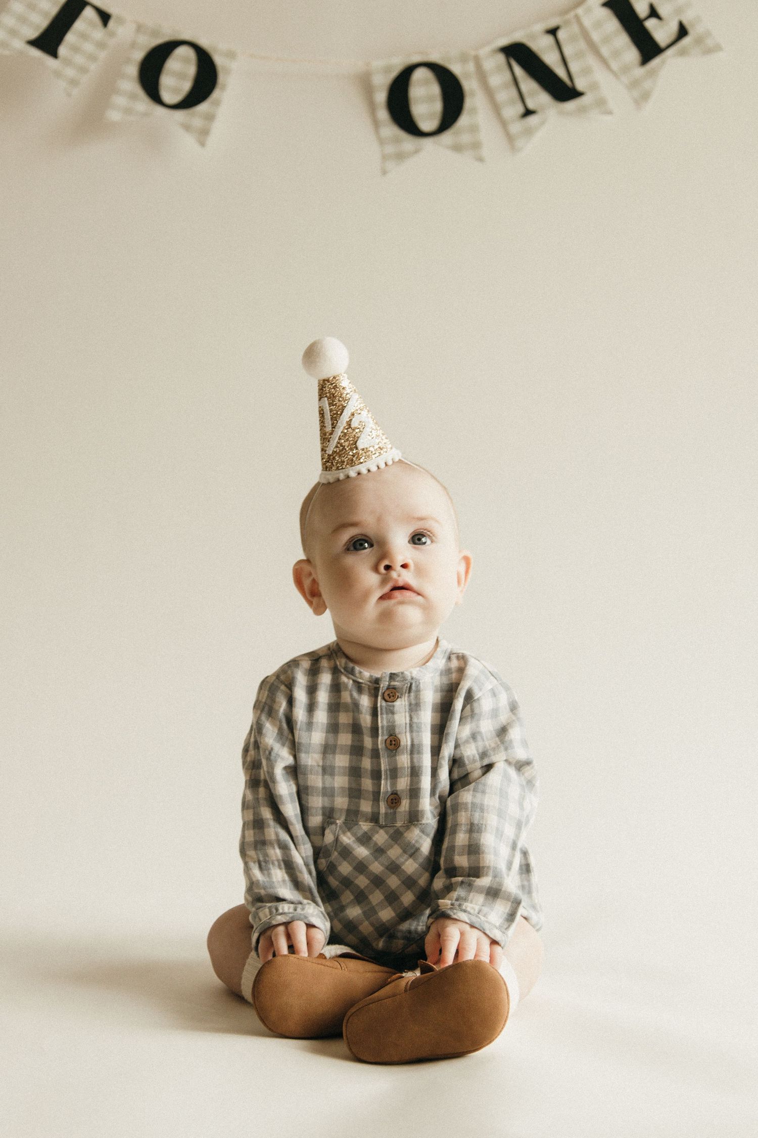 Baby's first birthday portrait with party hat, plaid romper, and 'Happy To One' banner.