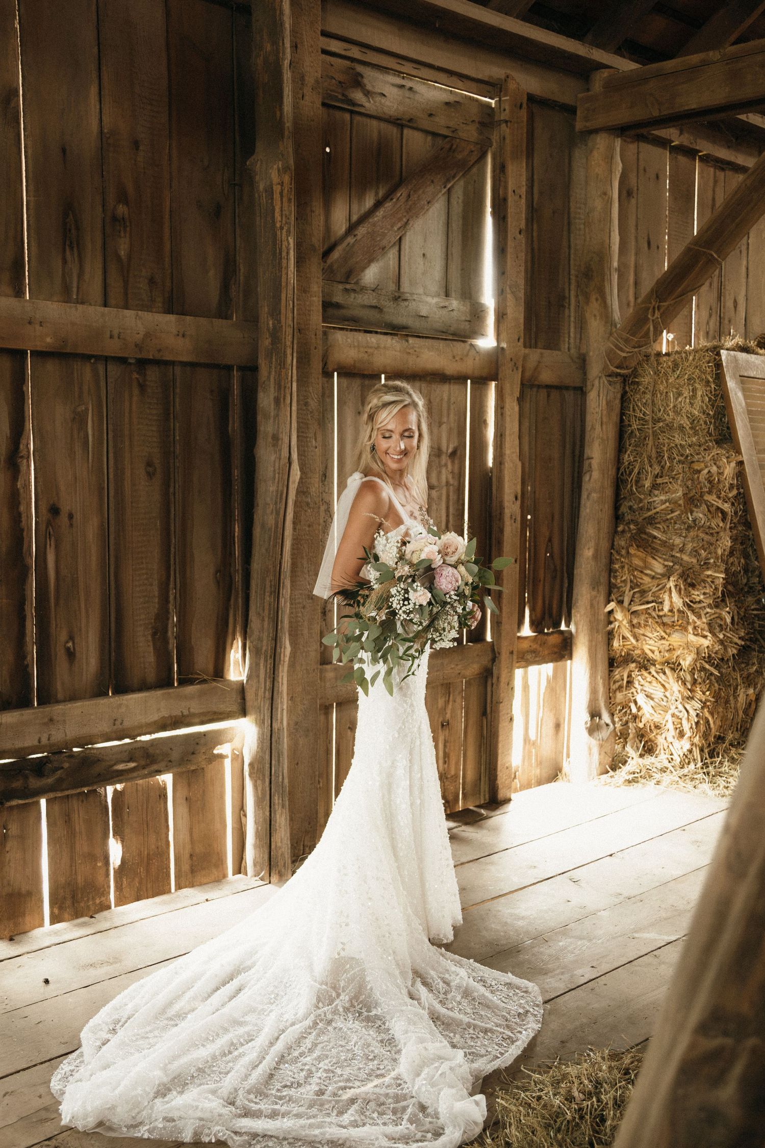 Bride in lace wedding gown holding floral bouquet inside rustic wooden barn with hay bales.