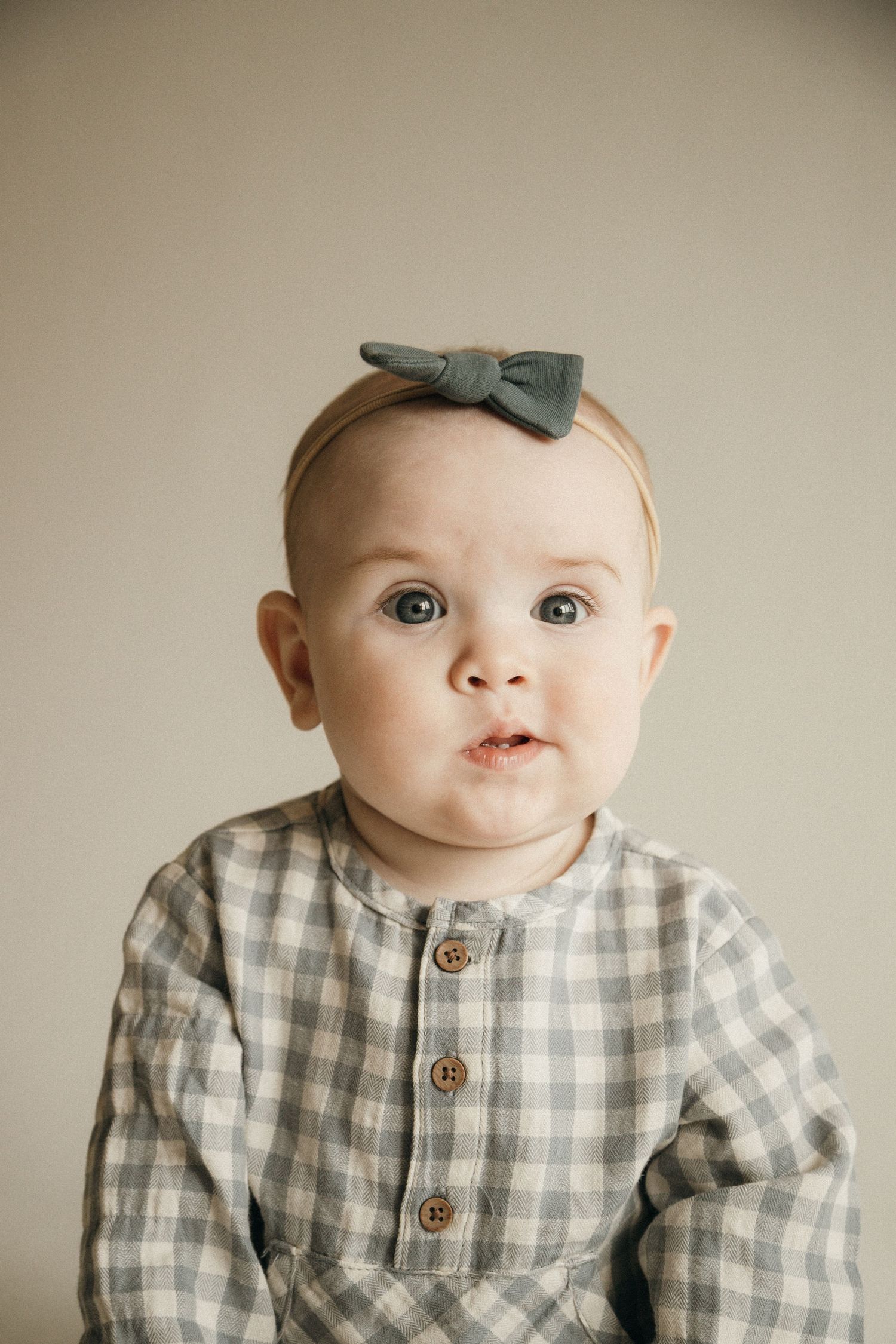 Adorable baby girl wearing a gray gingham outfit and bow headband looking curiously at camera.