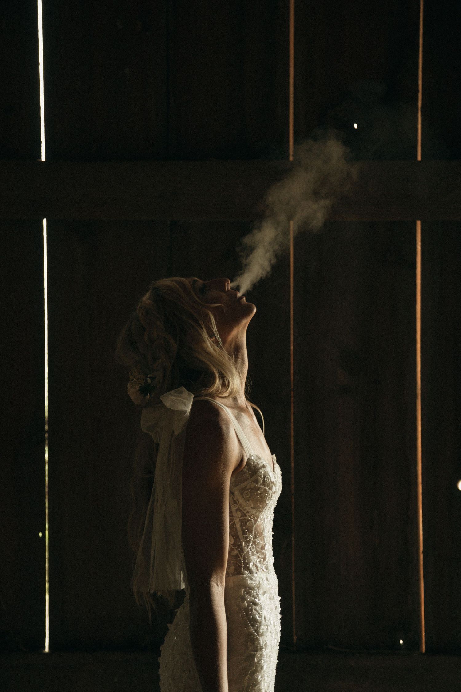 Bride in lace wedding dress exhaling smoke in dramatic barn lighting.