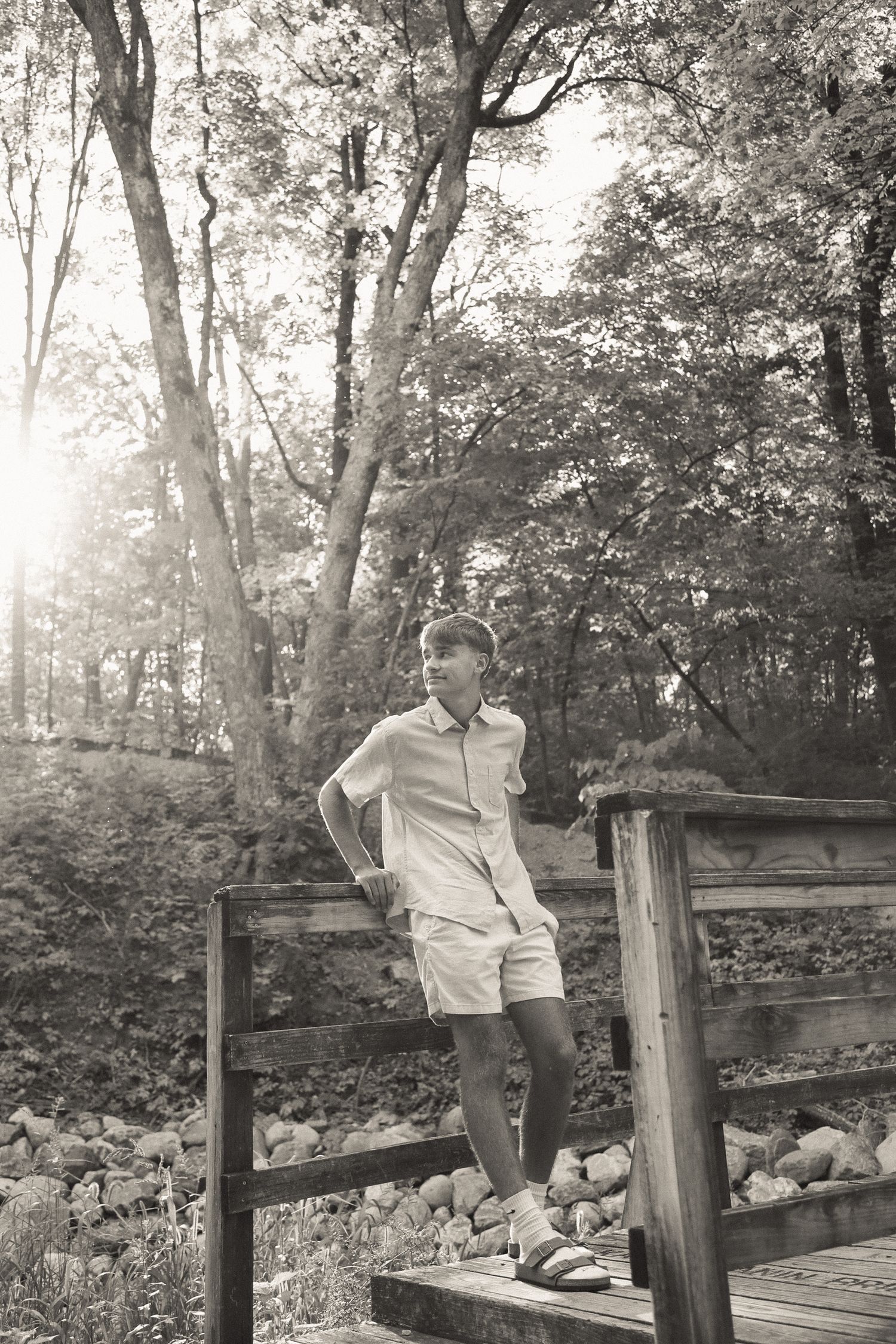 Black and white portrait of someone sitting on wooden fence near a pond.