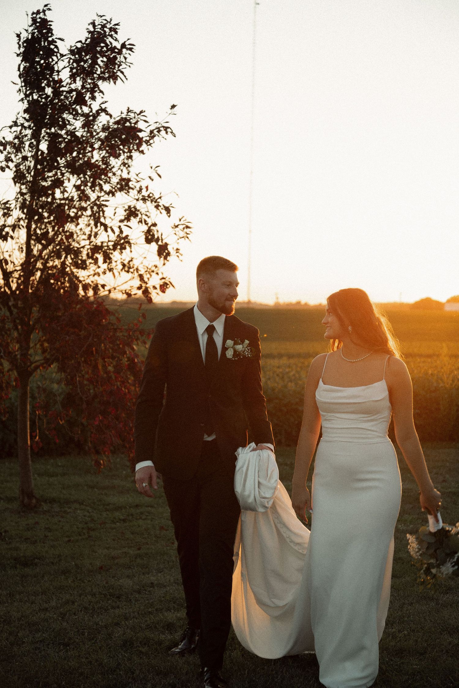 A couple in wedding attire walking together at sunset with a tree silhouetted in the background.