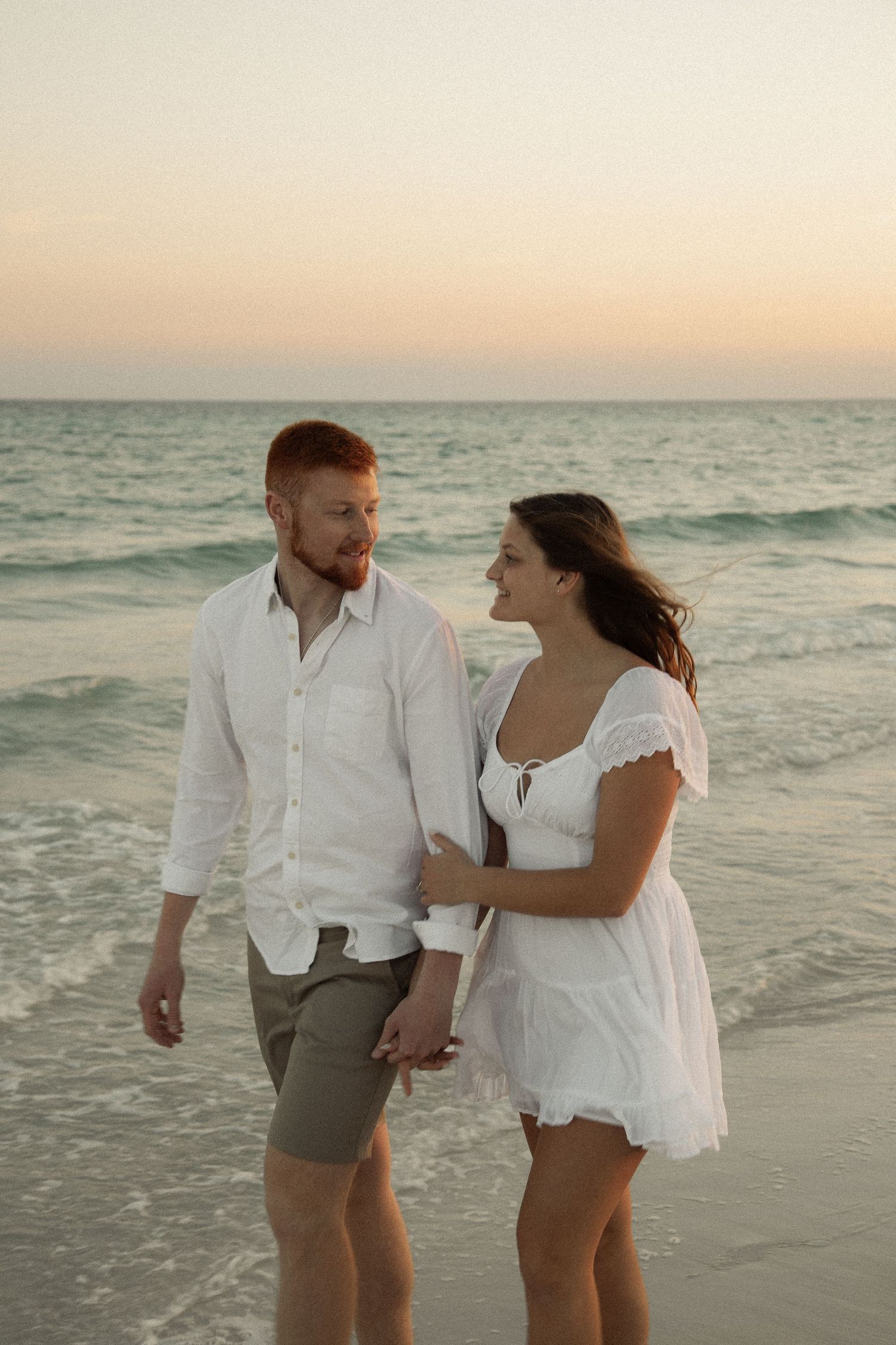 Couple in white outfits strolling along beach during sunset.